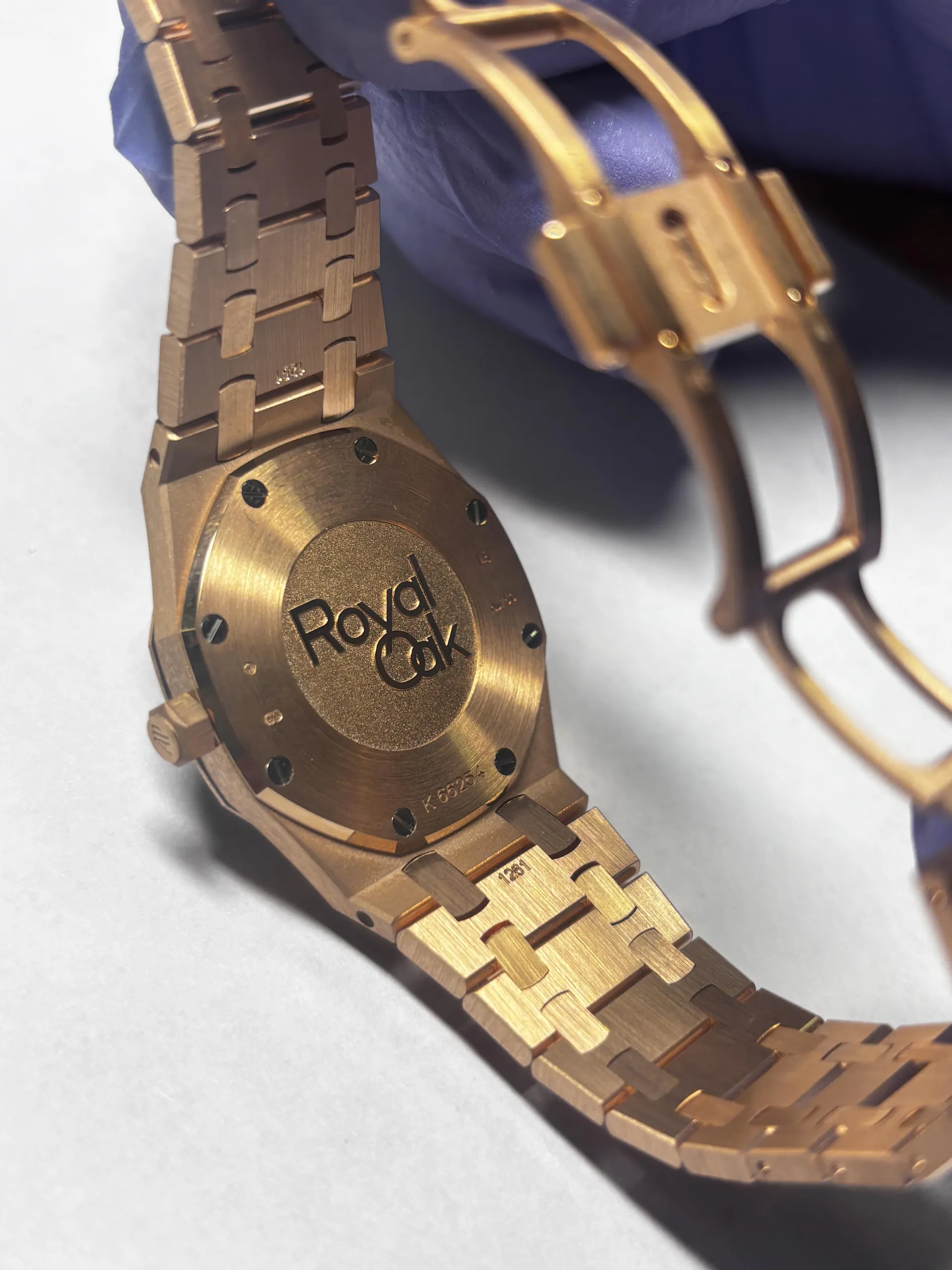 Zbliżenie na tylną część złotego zegarka z metalowym paskiem. Na tylnej pokrywie widoczny jest wygrawerowany napis 'Royal Oak' oraz oznaczenie 'K 585'. Zegarek trzymana jest w ręce w niebieskiej rękawiczce. Widoczne są również elementy zapięcia paska i śruby mocujące pokrywę.