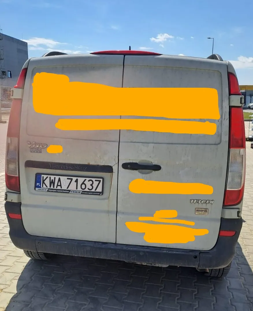 Tylny widok srebrnego vana marki Mercedes-Benz Vito. Na klapie bagażnika widoczna jest tablica rejestracyjna o numerze KWA 71637 oraz napisy 'Vito' i '113 CDI'. Część nadwozia jest zabrudzona, a niektóre elementy zostały zakryte żółtymi plamami. Pojazd stoi na kostce brukowej na tle nieba i budynku.