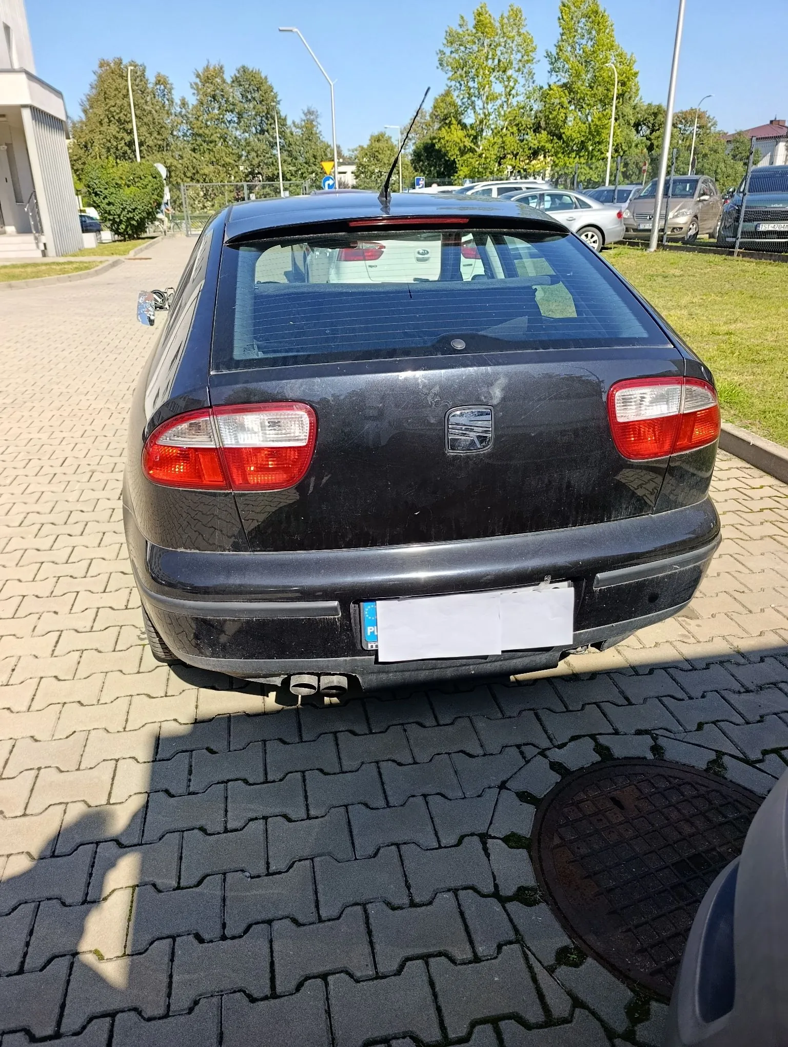 Tylny widok czarnego samochodu marki Seat zaparkowanego na kostce brukowej. Samochód ma czerwone tylne światła, centralnie umieszczone logo marki oraz zasłoniętą tablicę rejestracyjną z widocznym niebieskim paskiem Unii Europejskiej i literą PL. W tle widoczne są inne zaparkowane samochody, zielone drzewa oraz fragment budynku.