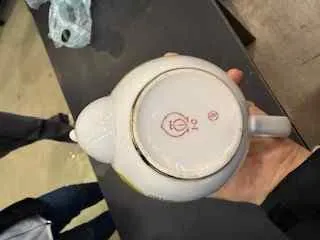 Biała ceramiczna czajniczek trzymany w dłoni, pokazujący dno z widocznym logo '1C2' i małym symbolem. W tle ciemny stół, fragment nogi osoby w ciemnych spodniach oraz niebieska torba plastikowa.