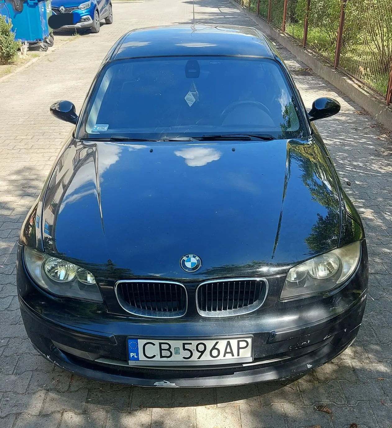 Czarny samochód marki BMW zaparkowany na kostce brukowej. Widok od przodu. Samochód ma polską tablicę rejestracyjną o numerze CB 596AP. W tle widoczna jest droga, ogrodzenie oraz fragment innego niebieskiego pojazdu.