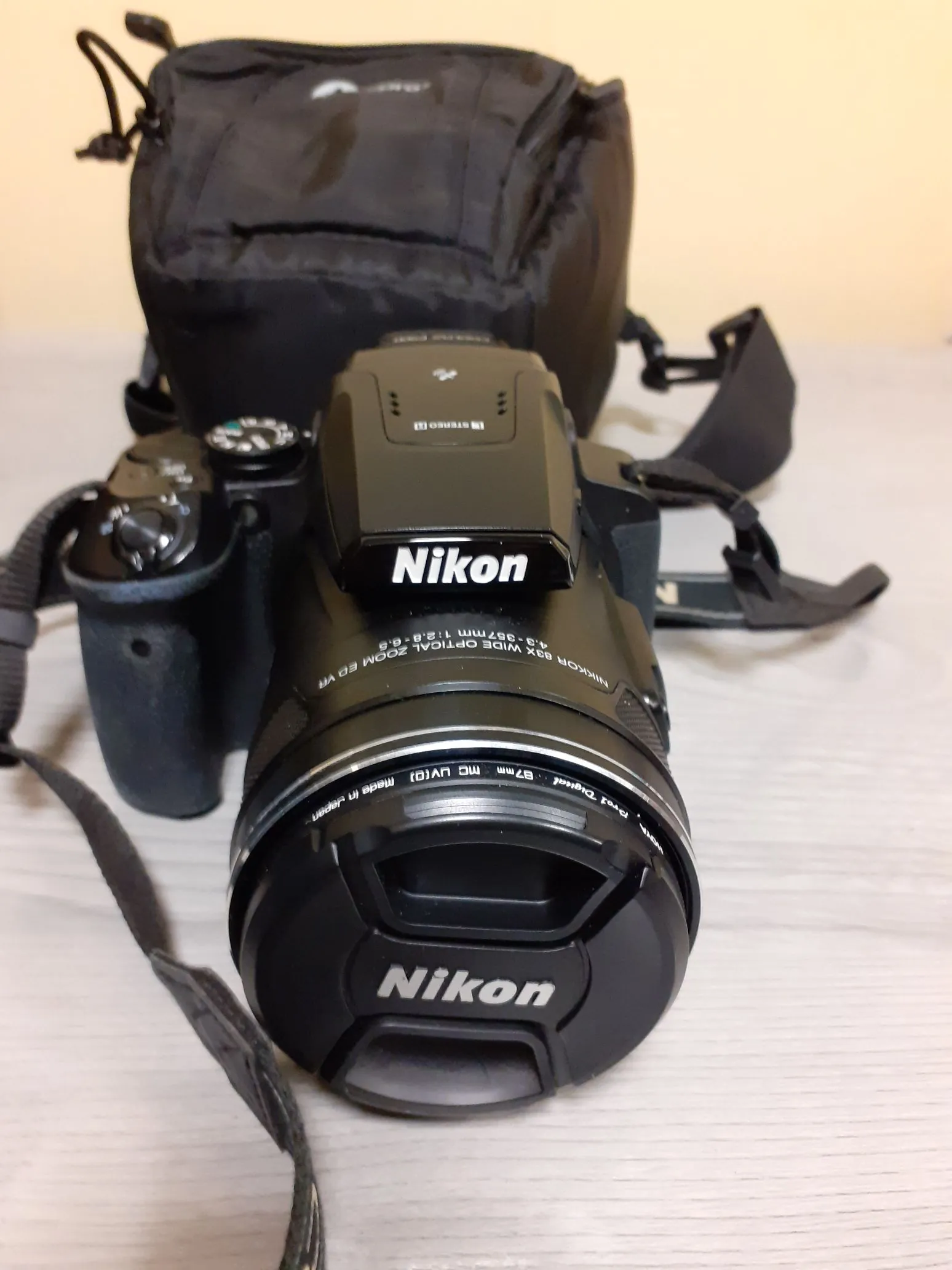 Czarny aparat fotograficzny marki Nikon z obiektywem, ustawiony przodem do widza. Na obiektywie widnieje napis 'Nikon' oraz specyfikacja techniczna: 'NIKKOR 83X WIDE OPTICAL ZOOM ED VR 4.9-357mm 1:2.8-6.6'. W tle znajduje się czarna torba fotograficzna oraz pasek aparatu.
