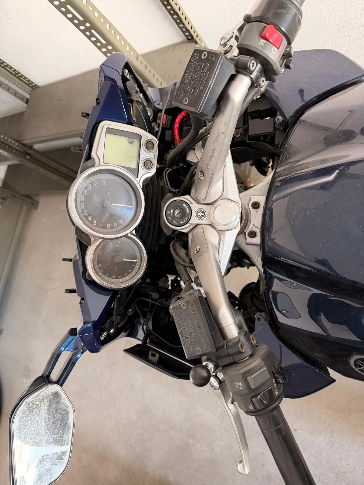 Widok z perspektywy kierowcy na kokpit ciemnoniebieskiego motocykla. Widoczne są analogowe zegary prędkości i obrotomierza, kierownica z manetkami oraz lusterko wsteczne po lewej stronie. Motocykl znajduje się w pomieszczeniu obok metalowego regału.