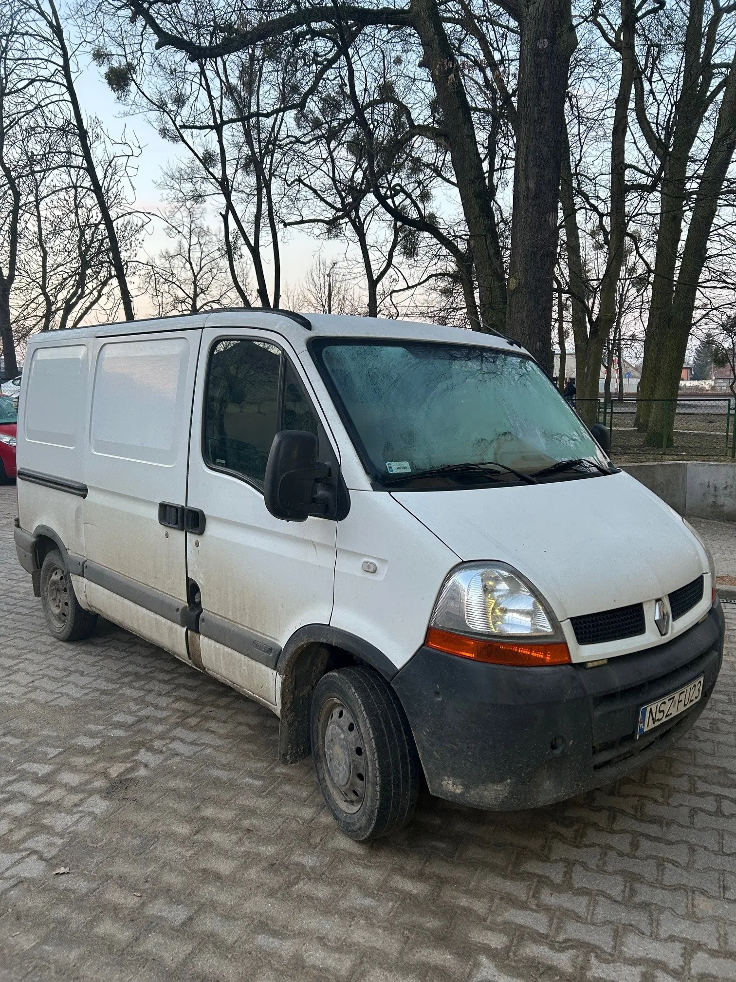 Biały dostawczy samochód marki Renault z tablicą rejestracyjną NSZ FU23 zaparkowany na brukowanej powierzchni. Samochód jest zabrudzony, zwłaszcza dolne części nadwozia. W tle widoczne są wysokie drzewa bez liści, fragment czerwonego samochodu po lewej stronie oraz ogrodzenie. Niebo jest jasne z lekkimi chmurami.