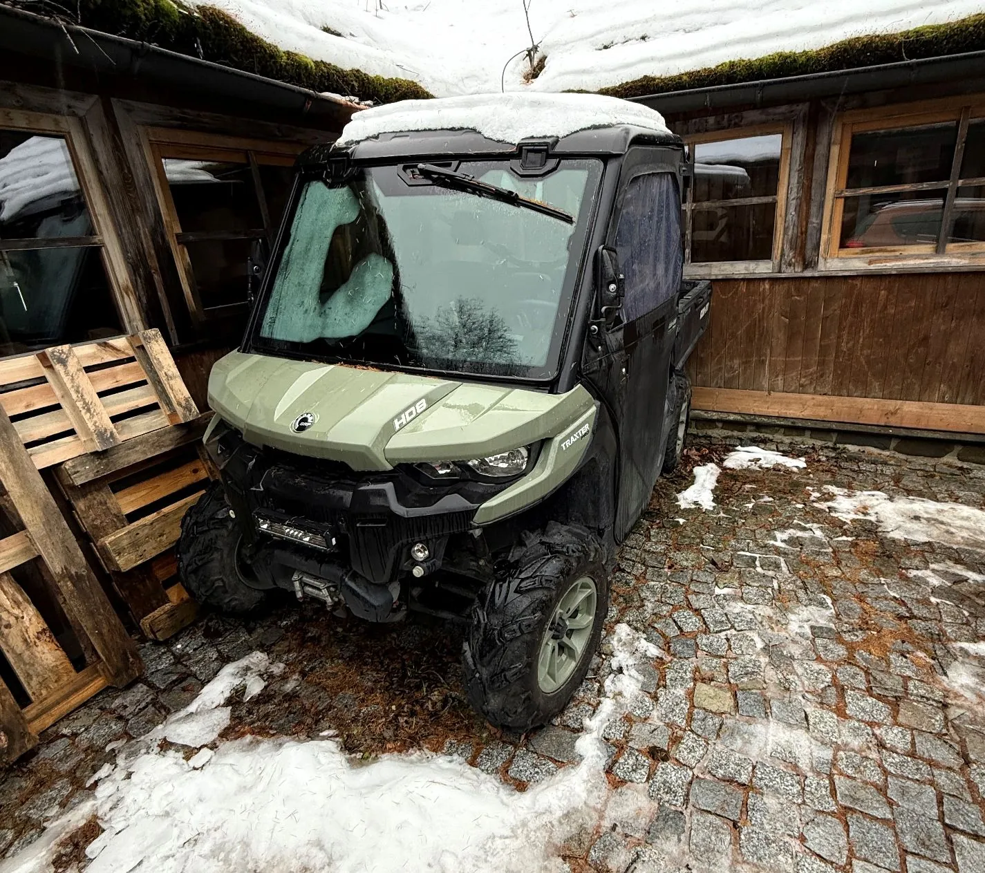 Oliwkowy pojazd typu UTV (Utility Task Vehicle) zaparkowany na brukowanym podjeździe przed drewnianym budynkiem. Na masce pojazdu widnieją napisy 'HOG' oraz 'TRAXTER'. Na dachu pojazdu oraz wokół niego leży śnieg. Po lewej stronie znajdują się ułożone drewniane palety.