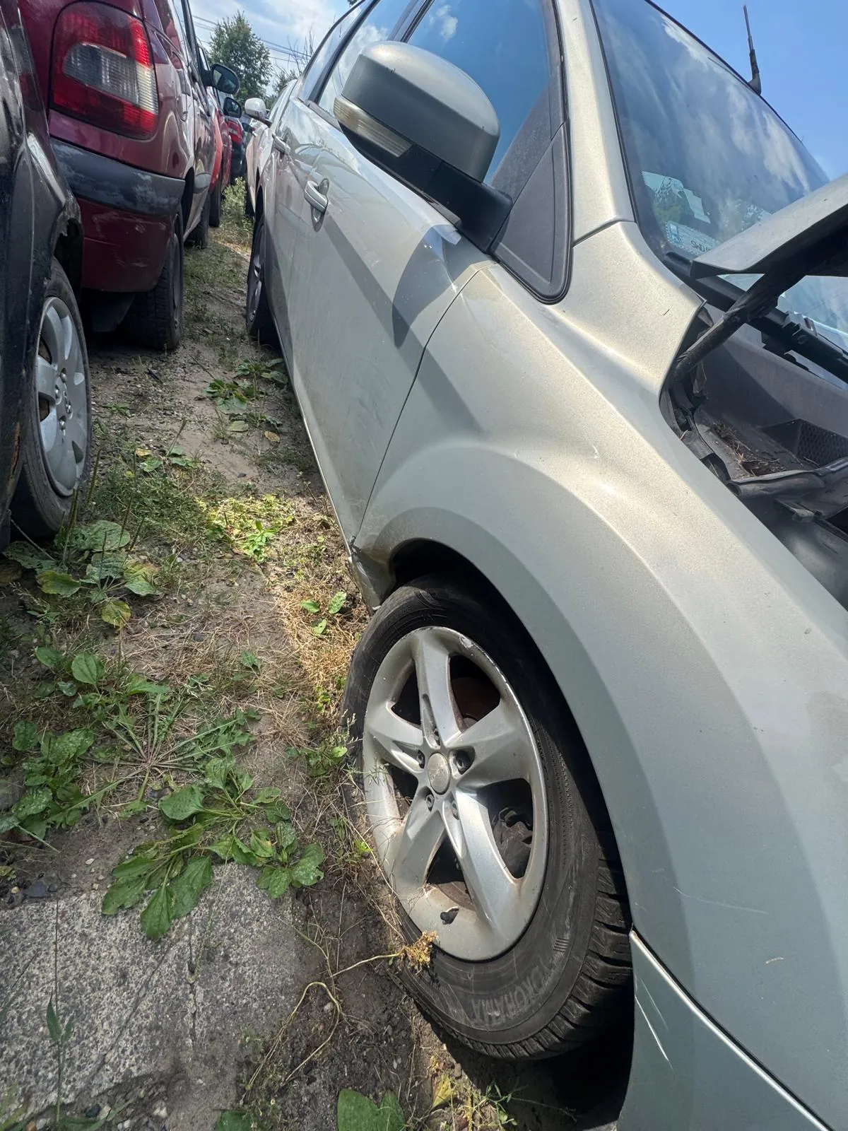 Zbliżenie na prawy przedni bok srebrnego samochodu zaparkowanego na nieutwardzonym terenie z trawą i chwastami. W tle widoczne są inne zaparkowane pojazdy, w tym czerwony samochód. Widoczna jest przednia opona z felgą aluminiową oraz fragment przedniej szyby i lusterka bocznego.