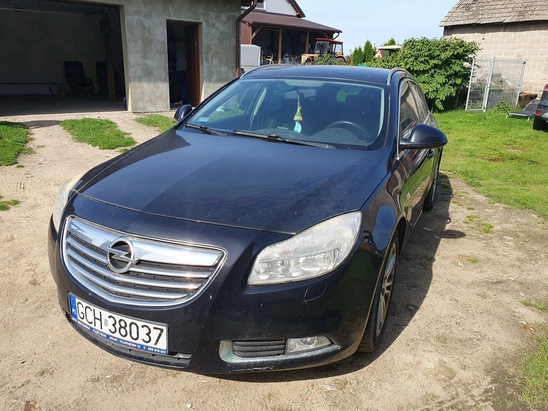 Czarny samochód osobowy marki Opel zaparkowany na piaszczystym podjeździe przed garażem. Pojazd ma polską tablicę rejestracyjną o numerze GCH 38037. W tle widoczny jest budynek garażowy, dom oraz zielona roślinność.