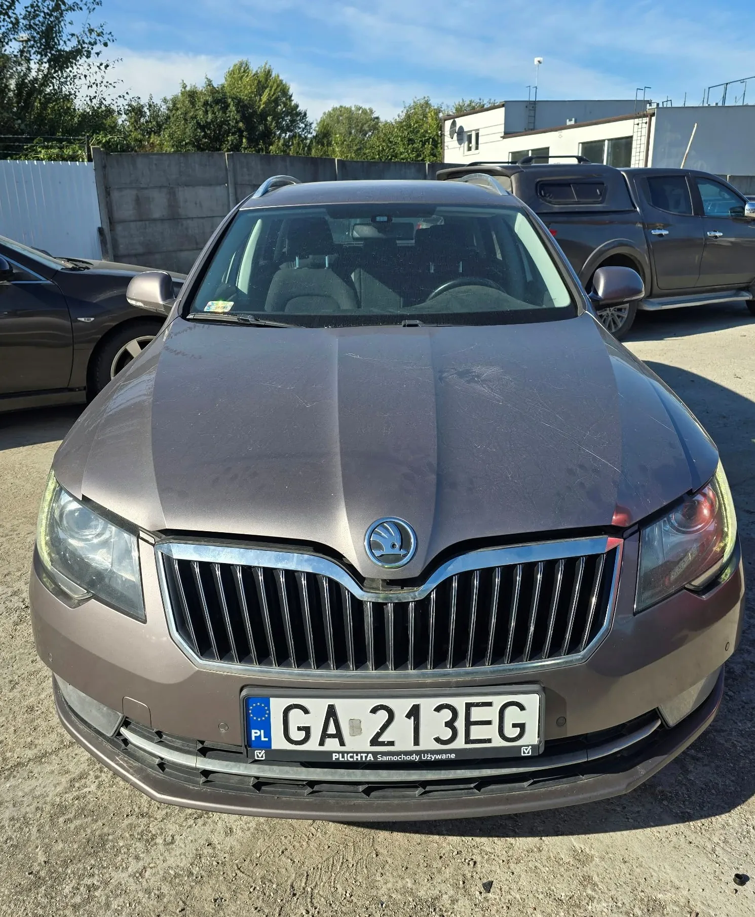 Widok z przodu brązowego samochodu marki Skoda zaparkowanego na betonowym placu. Pojazd ma tablicę rejestracyjną o numerze GA 213EG z oznaczeniem PL. Poniżej tablicy znajduje się napis 'PLICHTA Samochody Używane'. W tle widoczne są inne samochody, betonowy mur oraz biały budynek przemysłowy pod niebieskim niebem.