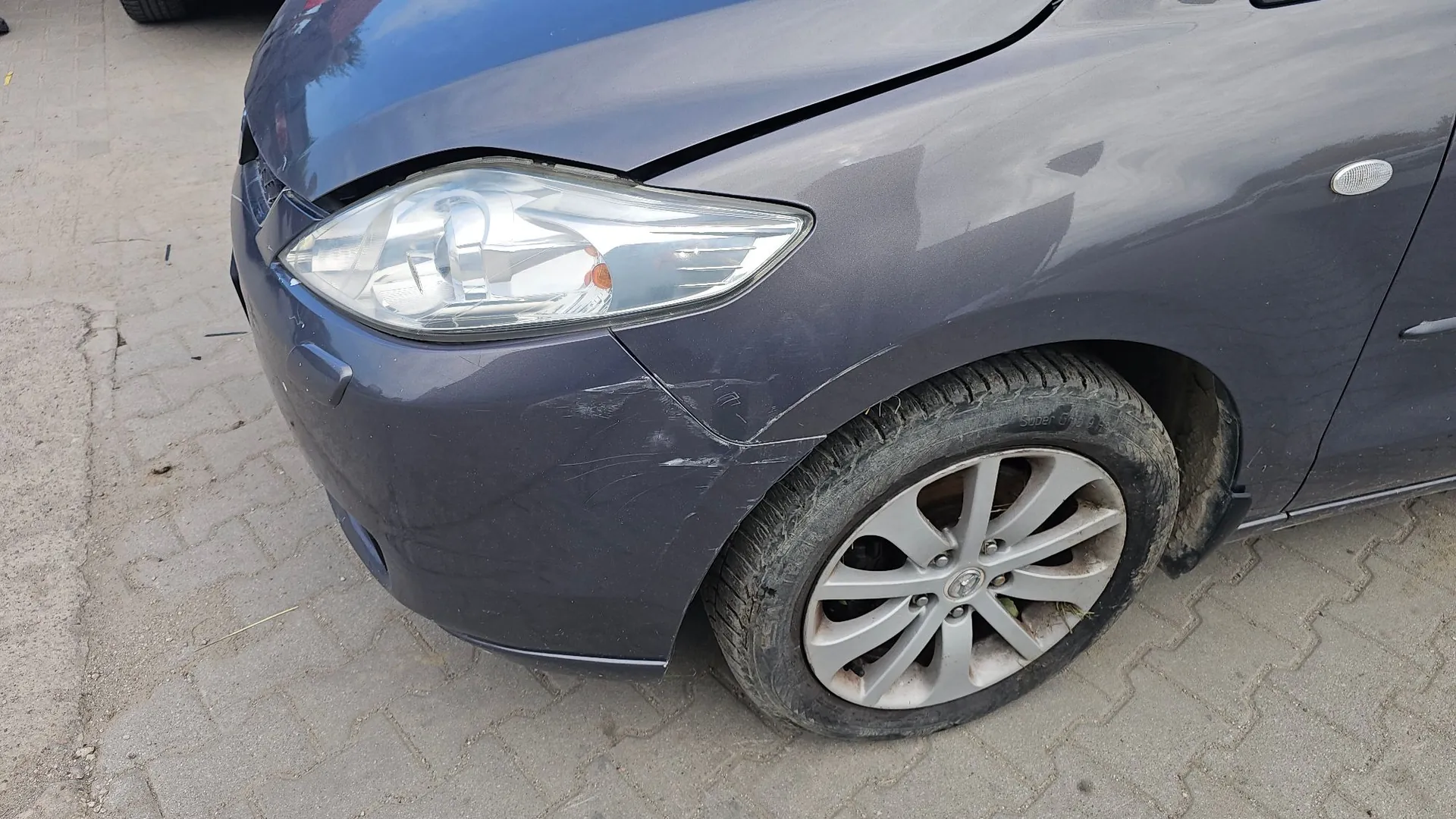 Zbliżenie na przedni lewy narożnik ciemnoniebieskiego samochodu. Widoczne są uszkodzenia karoserii: szczelina między maską a błotnikiem oraz liczne zarysowania i wgniecenia na przednim zderzaku w pobliżu reflektora. Samochód stoi na kostce brukowej.