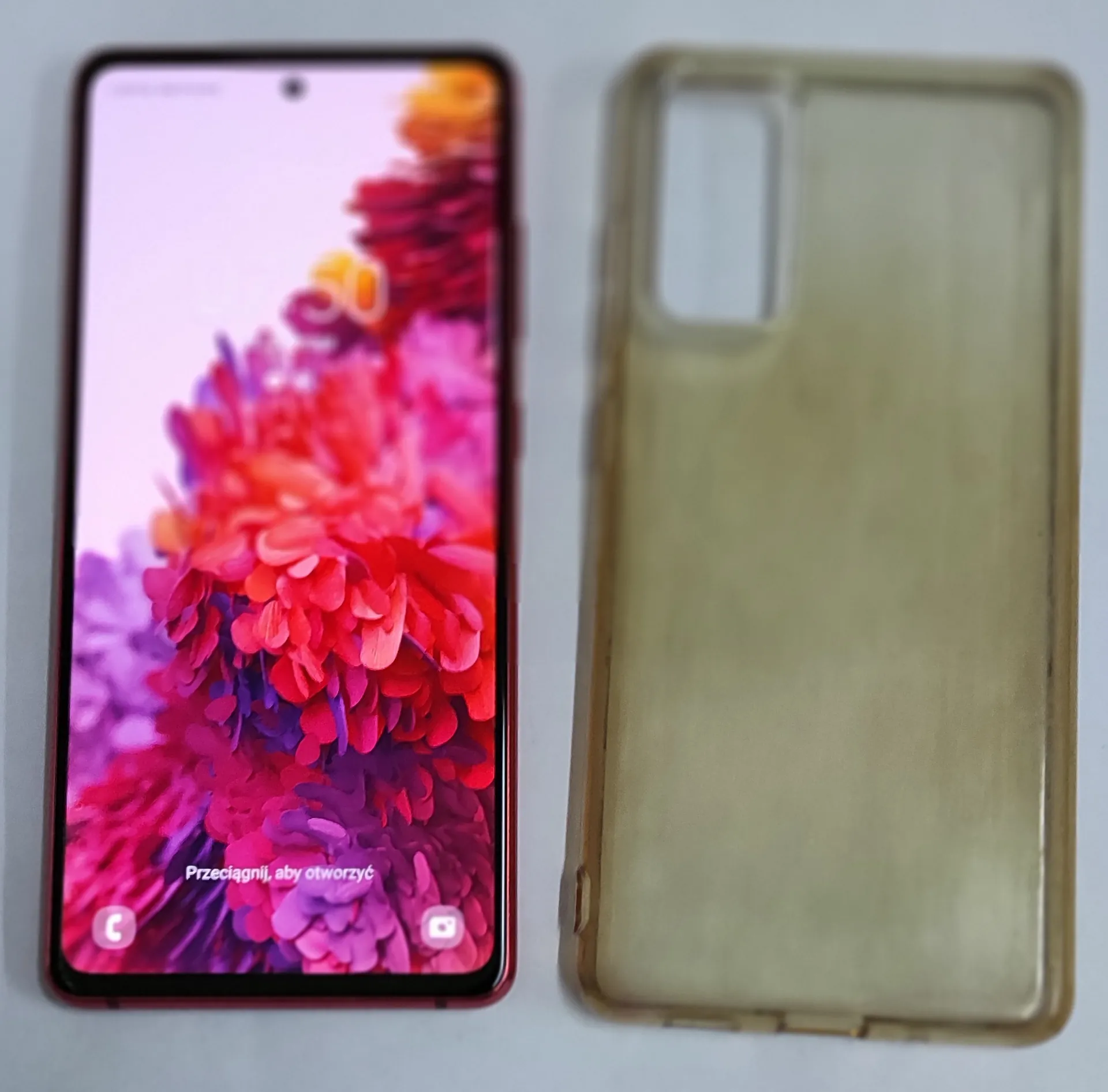 Obok siebie leżą dwa przedmioty: po lewej stronie smartfon z włączonym ekranem wyświetlającym kolorowe, czerwono-różowe kwiaty oraz napis na dole 'Przeciągnij, aby otworzyć'. Po prawej stronie znajduje się przezroczyste, lekko zżółknięte etui ochronne na telefon.