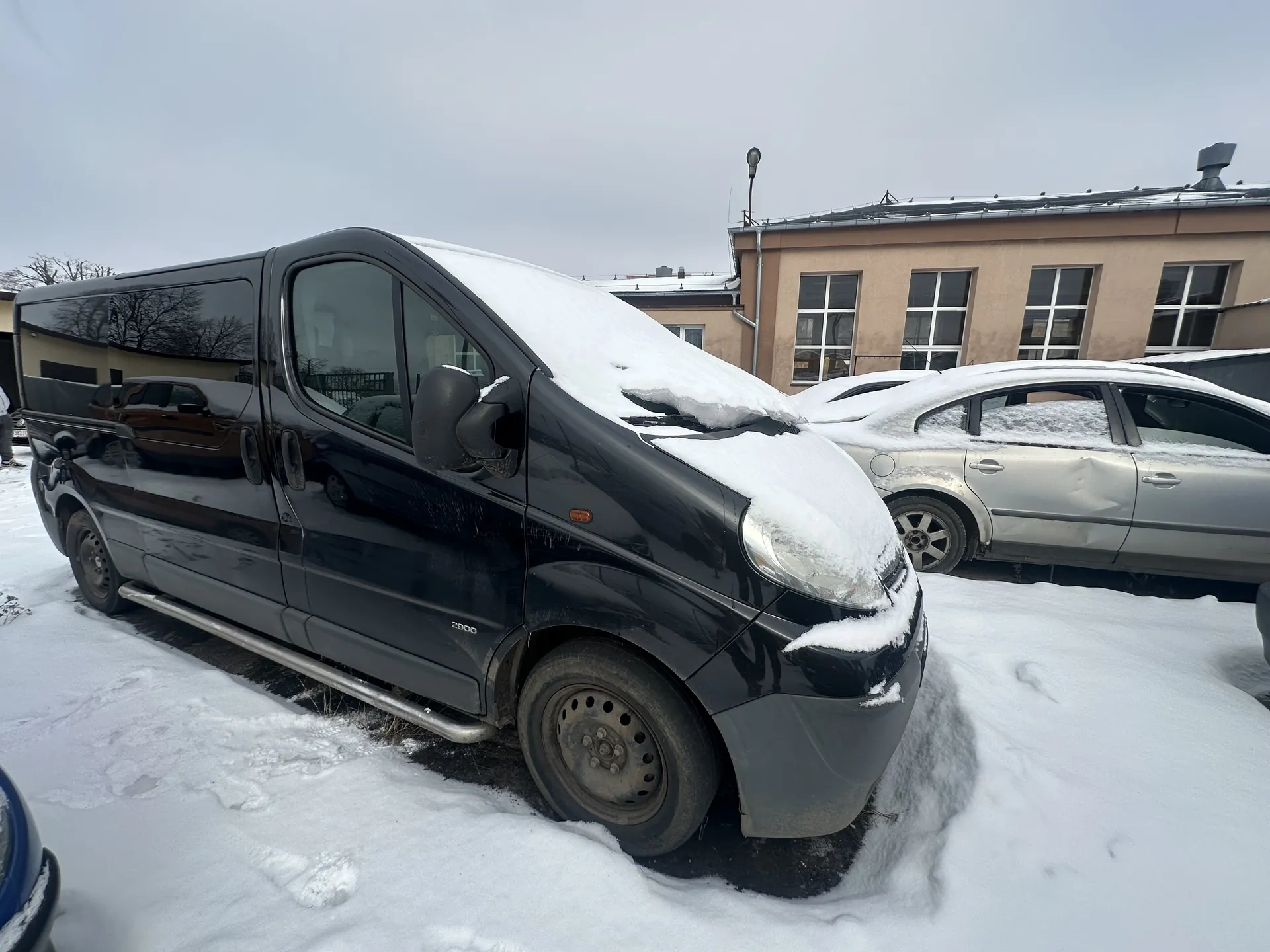 Czarny furgon z oznaczeniem 2900 na boku, pokryty śniegiem na dachu i masku, zaparkowany na śnieżnej powierzchni. Obok znajduje się srebrny samochód osobowy również z śniegiem na dachu. W tle budynki o beżowej elewacji z oknami oraz pochmurne niebo. Na ziemi widoczne ślady po stopionym śniegu.