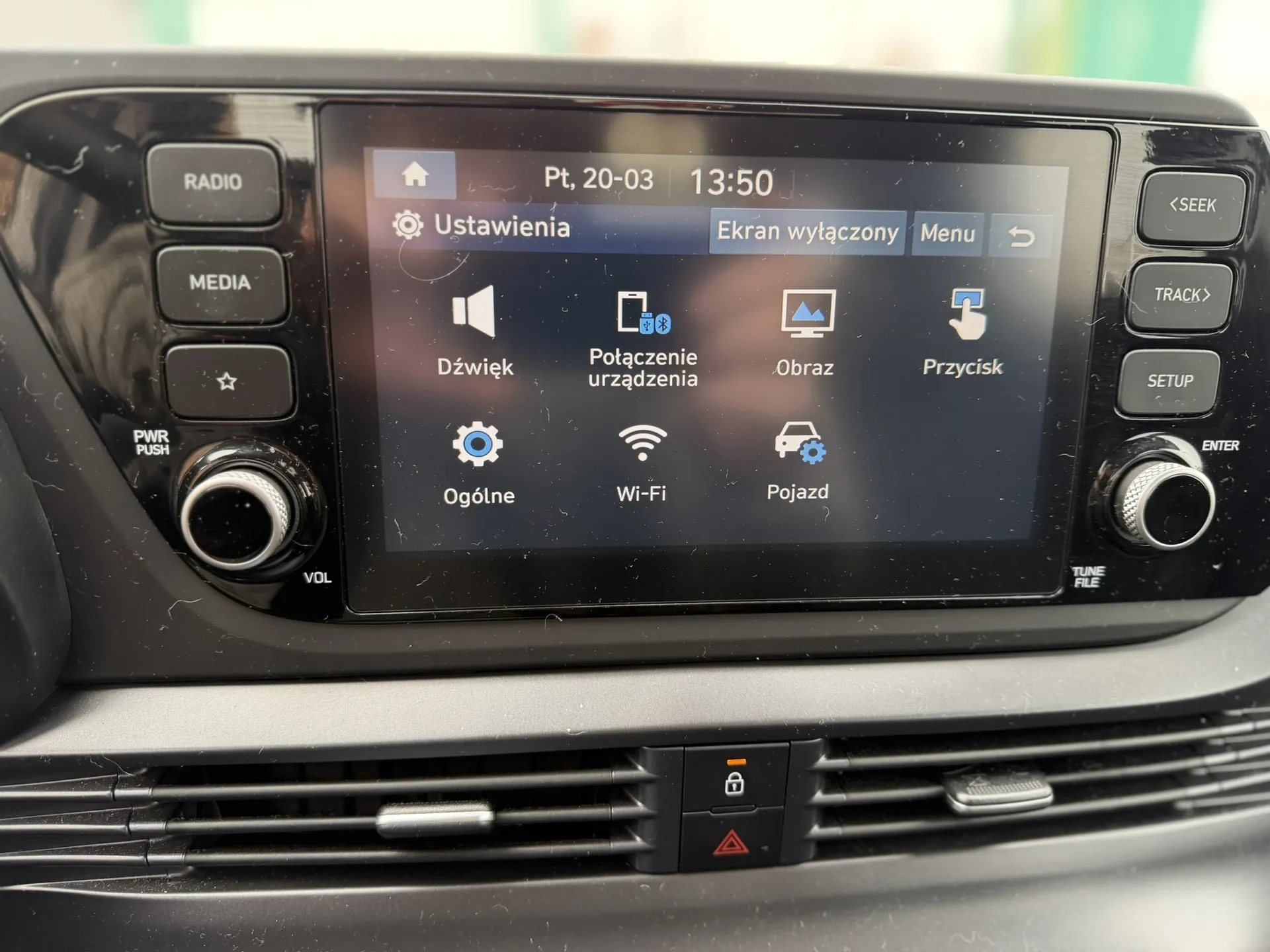 Zbliżenie na system rozrywki samochodowej z ekranem dotykowym. Na ekranie widoczny jest menu ustawień z napisem "Ustawienia", datą "Pt, 20-03", godziną "13:50" oraz opcjami: "Dźwięk", "Połączenie urządzenia", "Obraz", "Przycisk", "Ogólne", "Wi-Fi", "Pojazd". Po lewej stronie ekranu przyciski: "RADIO", "MEDIA", przycisk z ikoną gwiazdy, "PWR PUSH" i regulator głośności oznaczony jako "VOL". Po prawej stronie przyciski: "<SEEK", "TRACK>", "SETUP", "ENTER" oraz "TUNE FILE". Poniżej ekranu widoczne są klimatyzacyjne wywiewacze i przyciski ostrzegawcze z ikonami kłódki i trójkąta ostrzegawczego. Na ekranie również napisy "Ekran wyłączony" i "Menu".