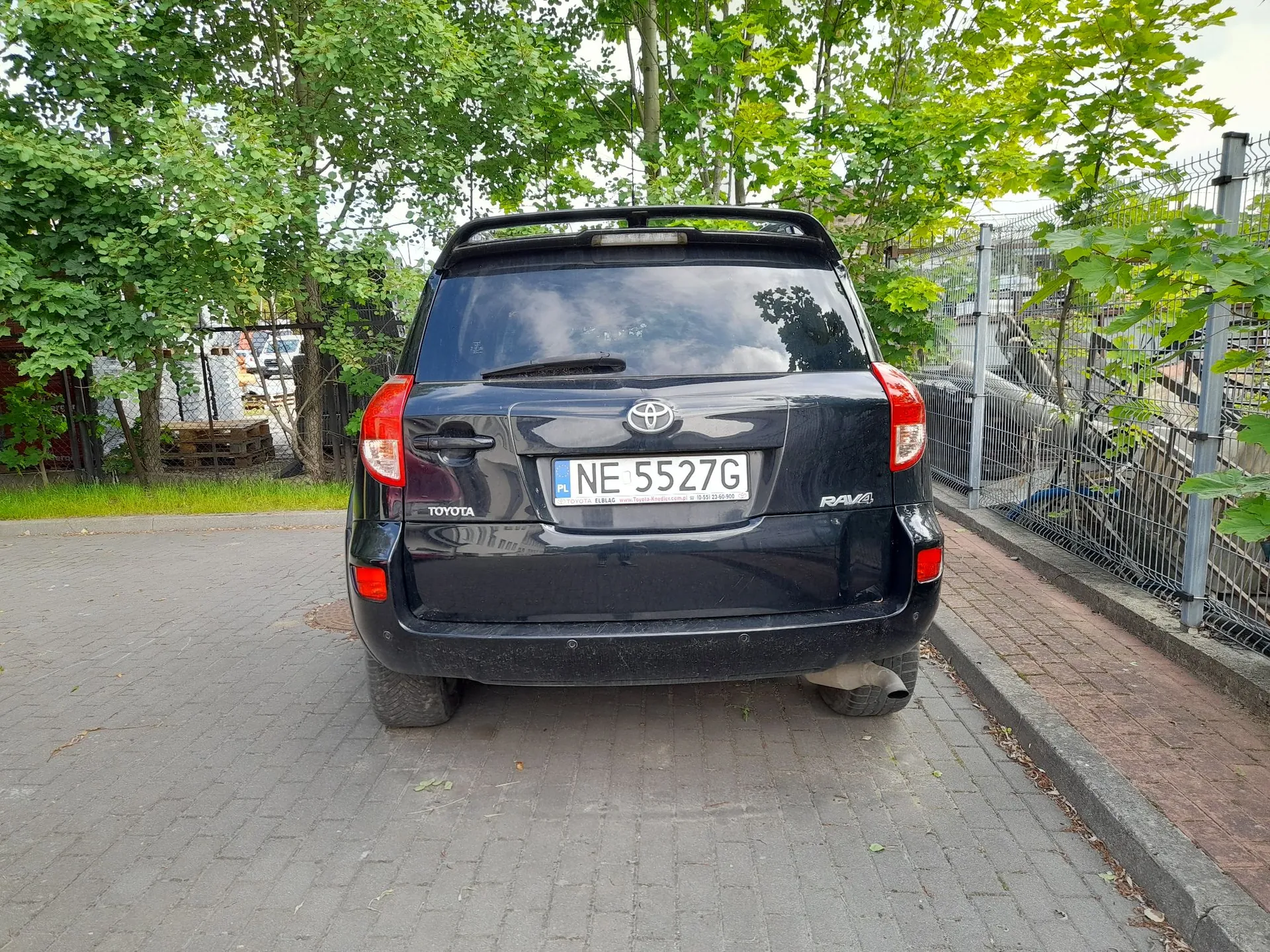 Tylny widok czarnego samochodu marki Toyota RAV4 zaparkowanego na kostce brukowej. Pojazd posiada polską tablicę rejestracyjną o numerze NE 5527G. W tle widoczne są zielone drzewa oraz metalowy ogrodzenie.