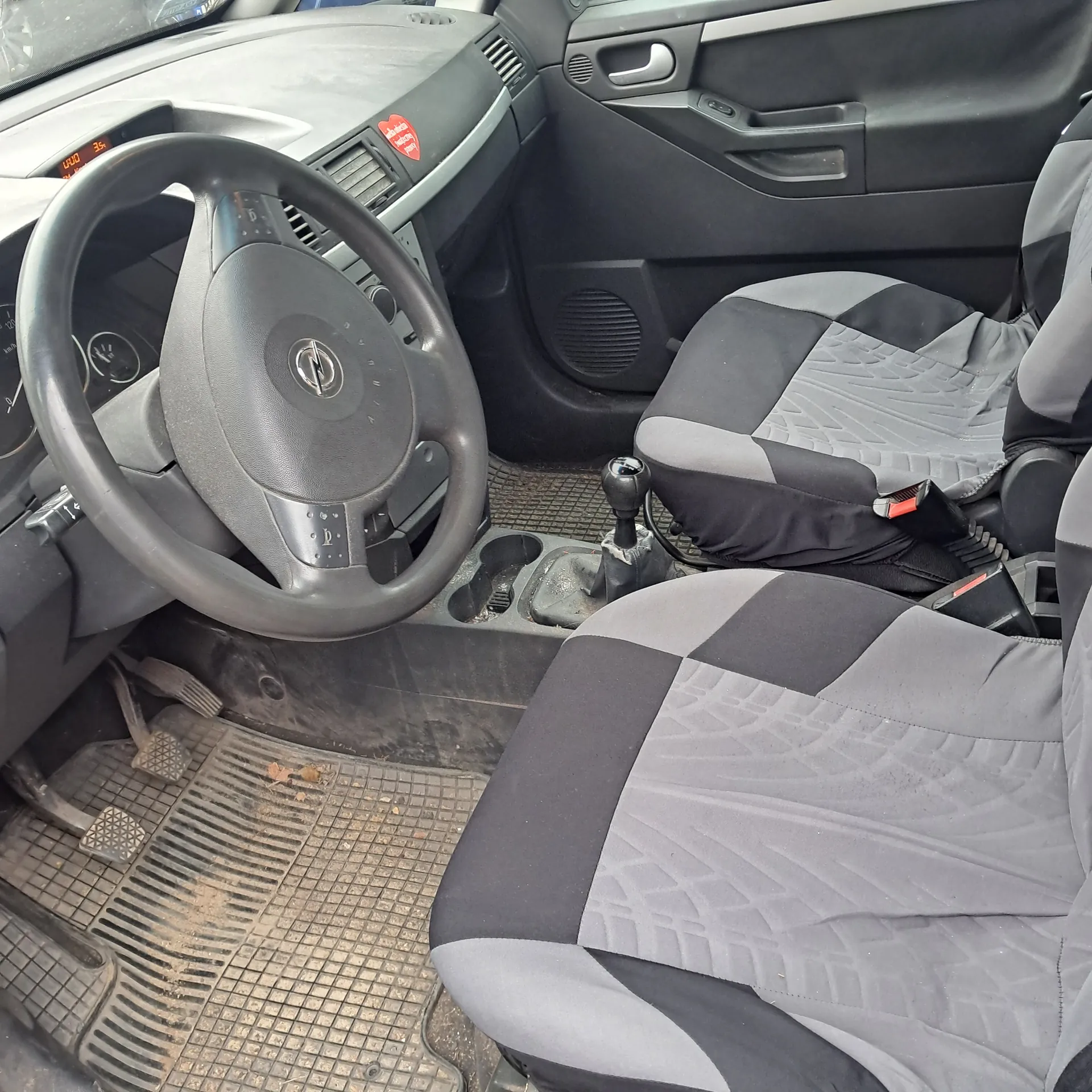 Wnętrze samochodu z kierownicą z logo Opel, przednimi siedzeniami w kolorze szaro-czarnym z geometrycznym wzorem, dźwignią zmiany biegów, deską rozdzielczą z czerwoną naklejką, brudnymi dywanikami podłogowymi oraz pedałami. Widoczny panel drzwiowy z uchwytem i głośnikiem.