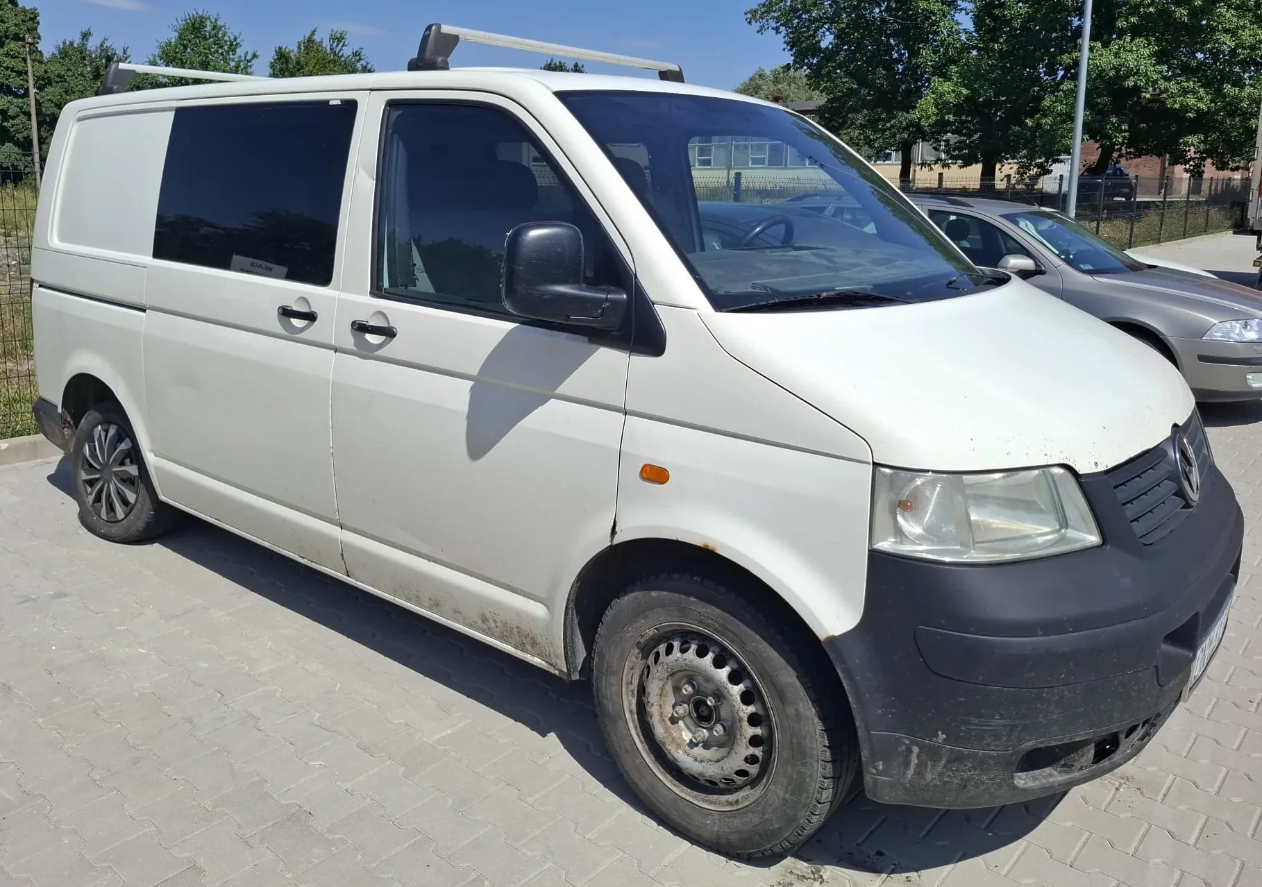 Biały samochód dostawczy marki Volkswagen Transporter zaparkowany na kostce brukowej. Pojazd posiada czarny zderzak przedni, czarne lusterka boczne oraz bagażnik dachowy. W tle widoczne są zielone drzewa, ogrodzenie oraz inny samochód w kolorze srebrnym.