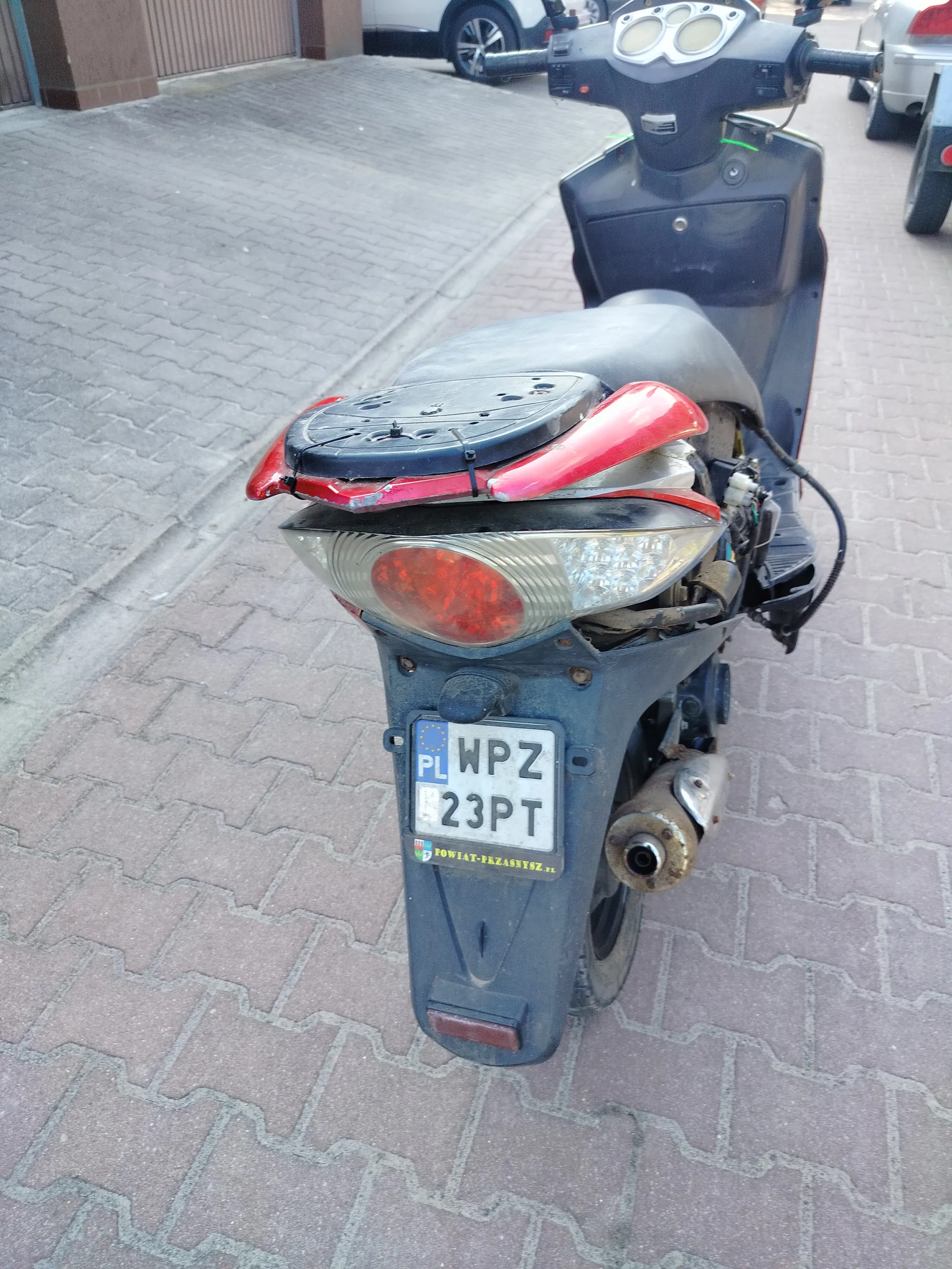 Zdjęcie przedstawia czarny skuter widziany od tyłu, zaparkowany na brukowanej powierzchni. Widoczny jest czerwony reflektor tylny, tablica rejestracyjna z napisem 'PL WPZ 23PT' oraz naklejka 'POWIAT-PRZASNYSZ.PL'. W tle znajdują się inne zaparkowane samochody i fragment budynku z brązową elewacją.