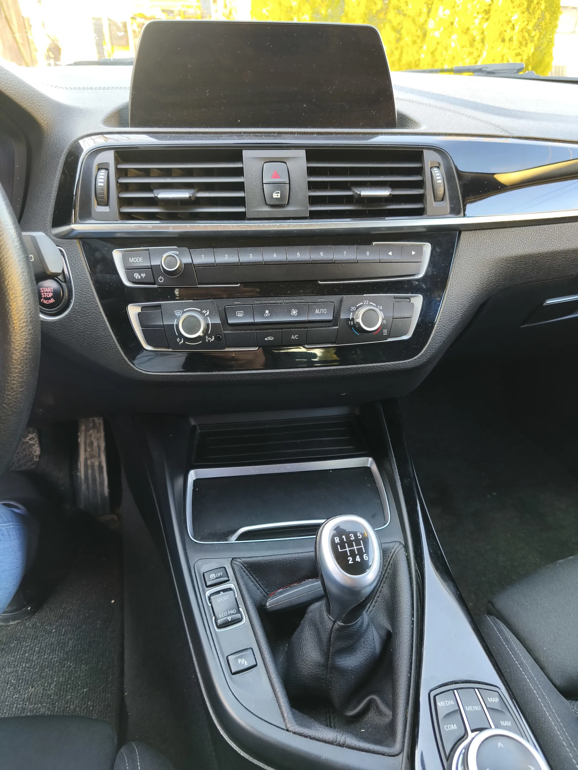 Wnętrze samochodu z widoczną konsolą środkową, kierownicą i przekładnią. Na konsoli środkowej znajdują się wentylacje, przycisk awaryjny z czerwonym trójkątem, ekran dotykowy, przyciski sterujące klimatyzacją (w tym napisy: MODE, AUTO, A/C) oraz pokrętła. Przekładnia manualna z oznaczeniem biegów R 1 3 5 / 2 4 6. Na kierownicy widoczny przycisk START STOP ENGINE. W tle przez przednią szybę widać żółte liście drzew. Wnętrze wykończone czarną tapicerką i plastikami.