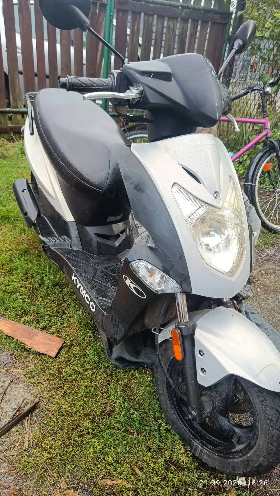 Srebrno-czarny skuter marki Kymco zaparkowany na trawie. Widoczny jest przód pojazdu z reflektorem, kierownicą i siedzeniem. Na boku skutera znajduje się napis 'KYMCO'. W tle widać drewniany płot oraz fragment różowego roweru.
