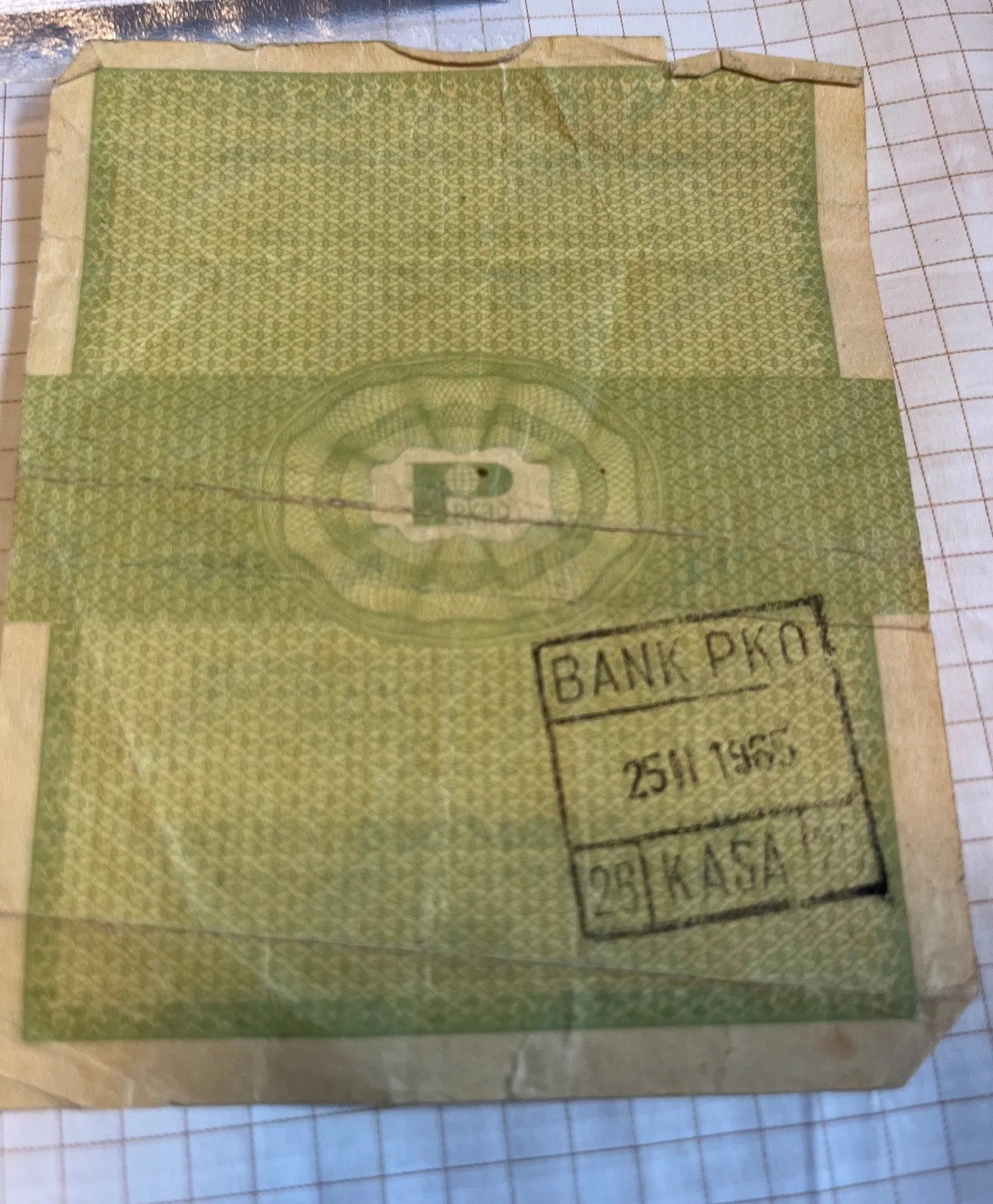 Zdjęcie starej, zniszczonej, zielonej koperty lub dokumentu z ozdobnym wzorem w tle. Na środku znajduje się jasny emblemat z literą 'P'. W prawym dolnym rogu widoczna jest czarna pieczątka prostokątna z tekstem: 'BANK PKO', poniżej data '25 II 1965' oraz napis '28 KASA'. Papier jest pognieciony i ma zagięcia, leży na białym papierze w kratkę.