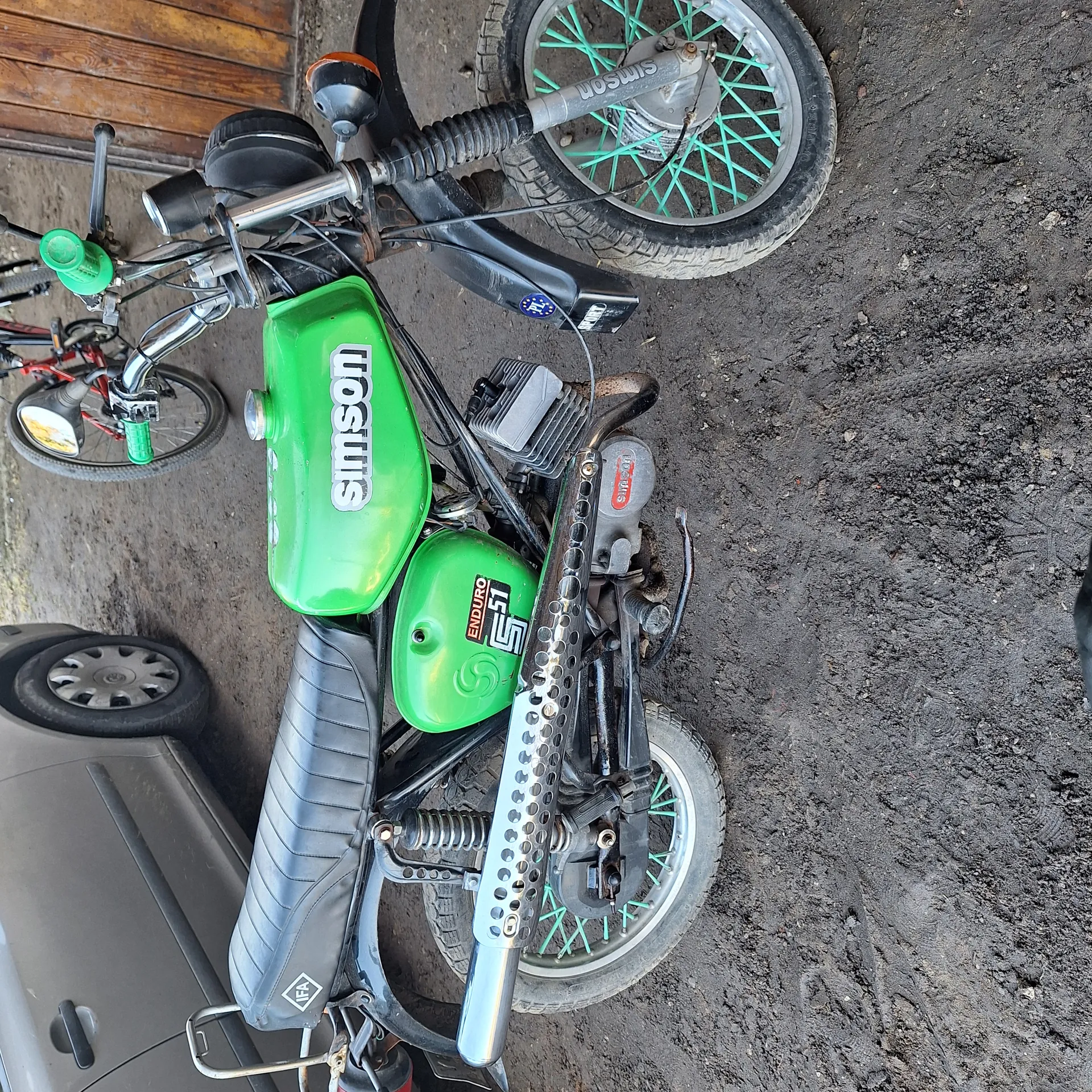 Zielony motocykl marki Simson leżący na boku na brudnej, ziemistej powierzchni. Widoczne zielone zbiorniki paliwa z napisem 'Simson' oraz 'Enduro 51' na jednym z nich. Koła z zielonymi promieniami, czarna siedziska i części mechaniczne. W tle fragment roweru, koło samochodu i drewniane deski.