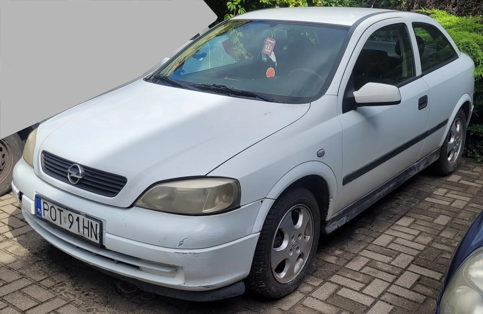Biały samochód osobowy marki Opel zaparkowany na kostce brukowej. Pojazd jest widoczny z przodu i z boku. Na przednim zderzaku znajduje się polska tablica rejestracyjna o numerze POT 91HN.