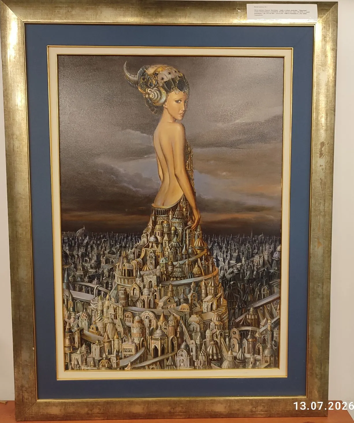 Obraz w złotej ramie z niebieskim passe-partout. Przedstawia on surrealistyczną postać kobiety, której dolna część ciała płynnie przechodzi w rozbudowane, fantastyczne miasto z licznymi wieżami, kopułami i mostami. Kobieta jest zwrócona tyłem, spogląda przez ramię w stronę widza i ma na głowie ozdobny, złoty hełm z rogiem. Tło stanowi ciemne, zachmurzone niebo. W prawym dolnym rogu zdjęcia widnieje data 13.07.2026.