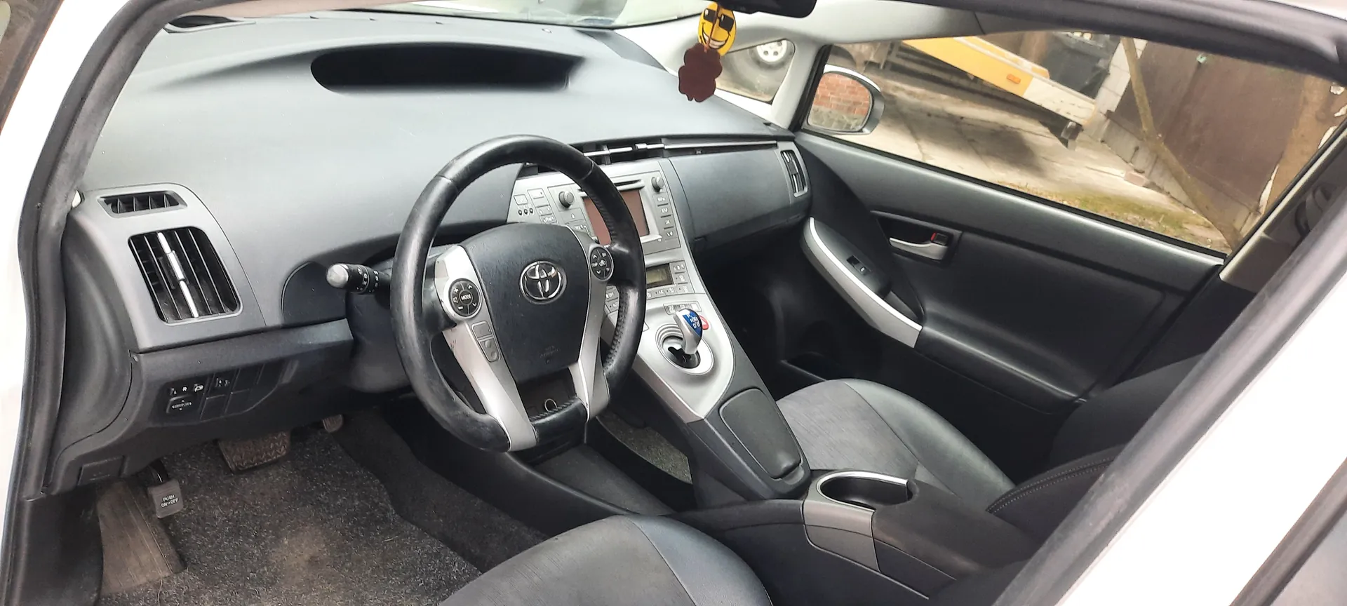 Wnętrze samochodu marki Toyota z widocznym kierownicą z logo, deska rozdzielcza z wentylacjami, centralną konsolą z przyciskami i dźwignią biegów. Siedzenia w kolorze szaro-czarnym, otwarte drzwi po stronie kierowcy. W lusterku wstecznym wiszą żółty uśmiechnięty emoji i brązowa ozdoba. Za oknem widoczny żółty pojazd i ceglana ściana.