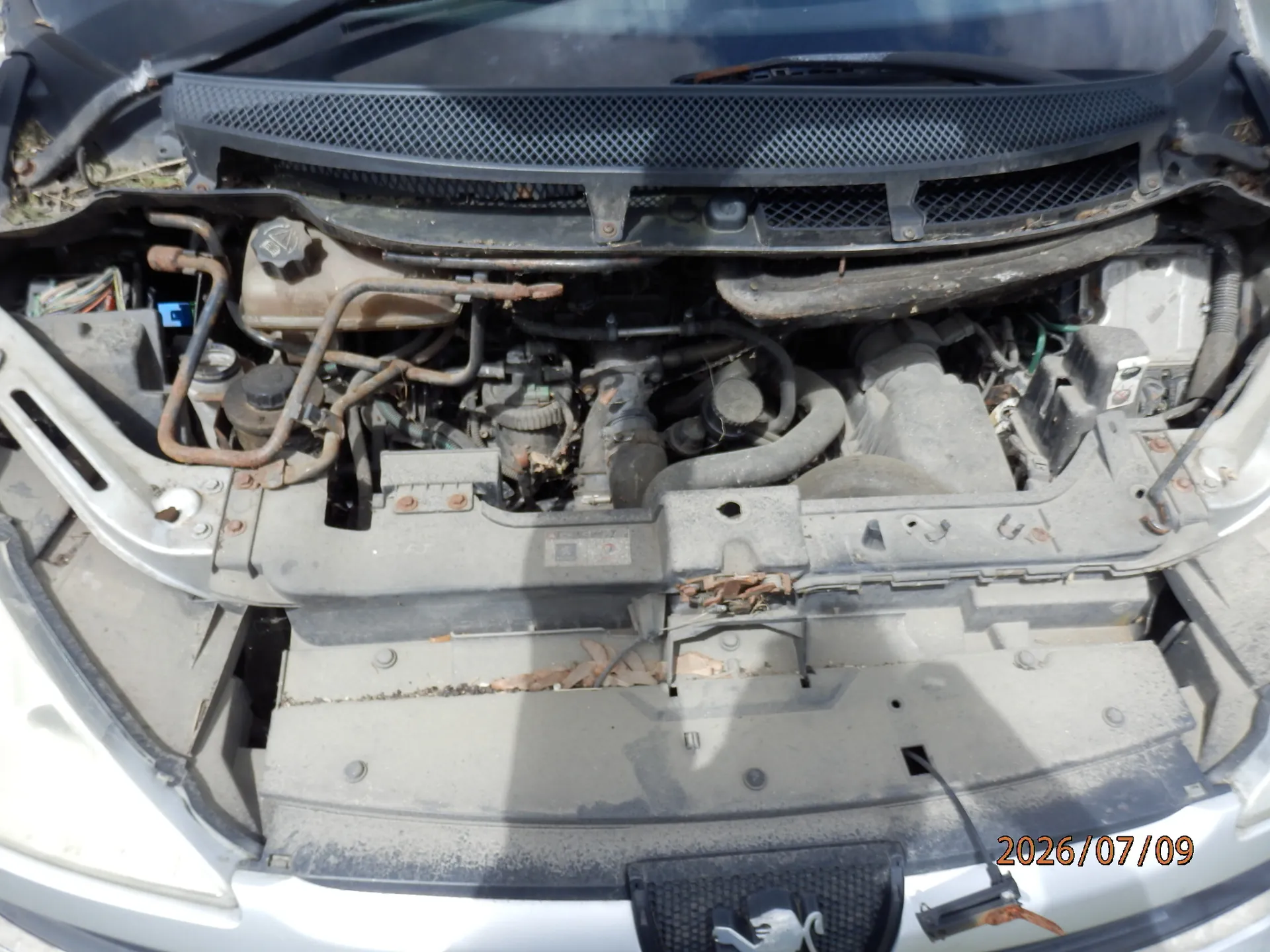 Widok z góry na otwartą komorę silnika samochodu marki Peugeot. Widoczne są elementy mechaniczne, przewody, zbiornik płynu oraz zakurzone powierzchnie plastikowe. W prawym dolnym rogu znajduje się data 2026/07/09.