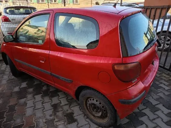 Czerwony samochód osobowy typu hatchback zaparkowany na brukowanej powierzchni. Widok z tyłu i boku. Samochód ma zabrudzoną karoserię, brak kołpaka na tylnej oponie oraz rdzewną felgę. W tle metalowe ogrodzenie i częściowo widoczne inne pojazdy. Na szybach odbijają się budynki.