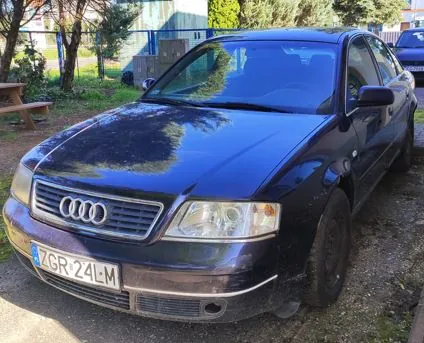 Granatowy samochód marki Audi zaparkowany na podjeździe. Widoczny jest przód pojazdu z charakterystycznym grillem z czterema pierścieniami oraz tablicą rejestracyjną o numerze ZGR 24L M. W tle widać zieleń, drzewa oraz fragment niebieskiego ogrodzenia.