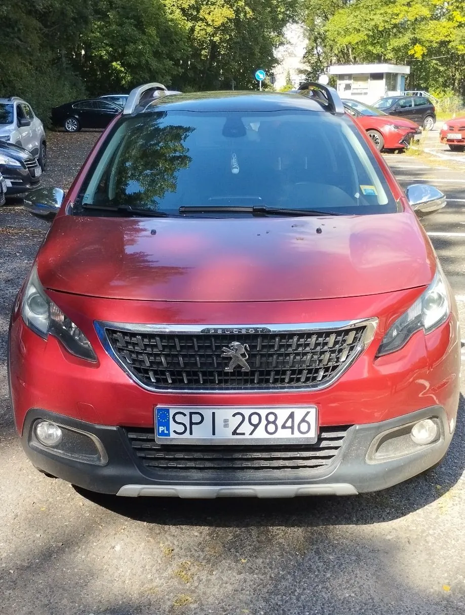 Widok z przodu czerwonego samochodu marki Peugeot zaparkowanego na zewnątrz. Pojazd ma polską tablicę rejestracyjną o numerze SPI 29846. W tle widoczne są inne zaparkowane samochody oraz zielone drzewa.