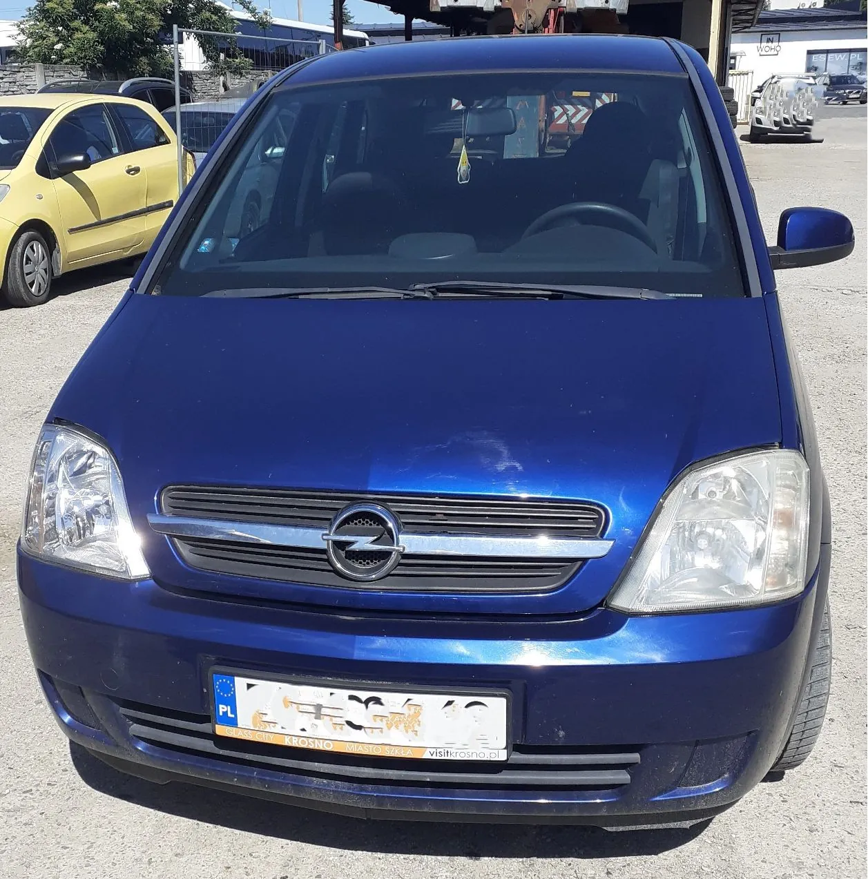 Niebieski samochód marki Opel zaparkowany na zewnątrz. Widok z przodu, ukazujący maskę, reflektory, grill z logo producenta oraz tablicę rejestracyjną z oznaczeniem PL. W tle widoczny jest fragment żółtego samochodu oraz budynki przemysłowe.