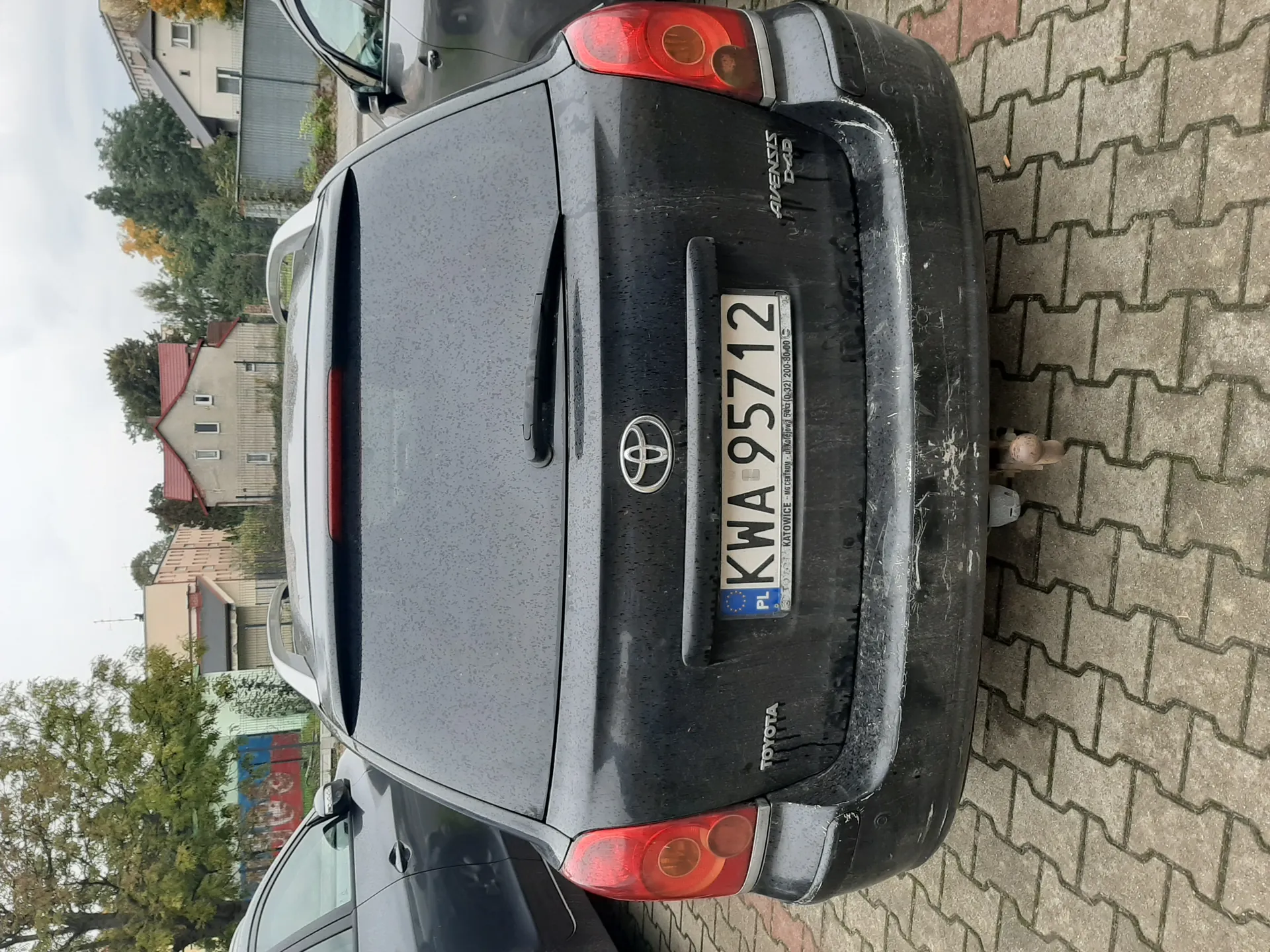 Tył czarnego samochodu Toyota Avensis D-4D z tablicą rejestracyjną KWA 95712 (PL). Samochód jest zaparkowany na brukowanej powierzchni, pokryty kroplami wody i zabrudzony. W tle widoczne są inne pojazdy, drzewa, budynki mieszkalne oraz ogrodzenie. Na tablicy rejestracyjnej widoczny napis "KATOWICE" oraz dodatkowe informacje. Niebo jest zachmurzone.