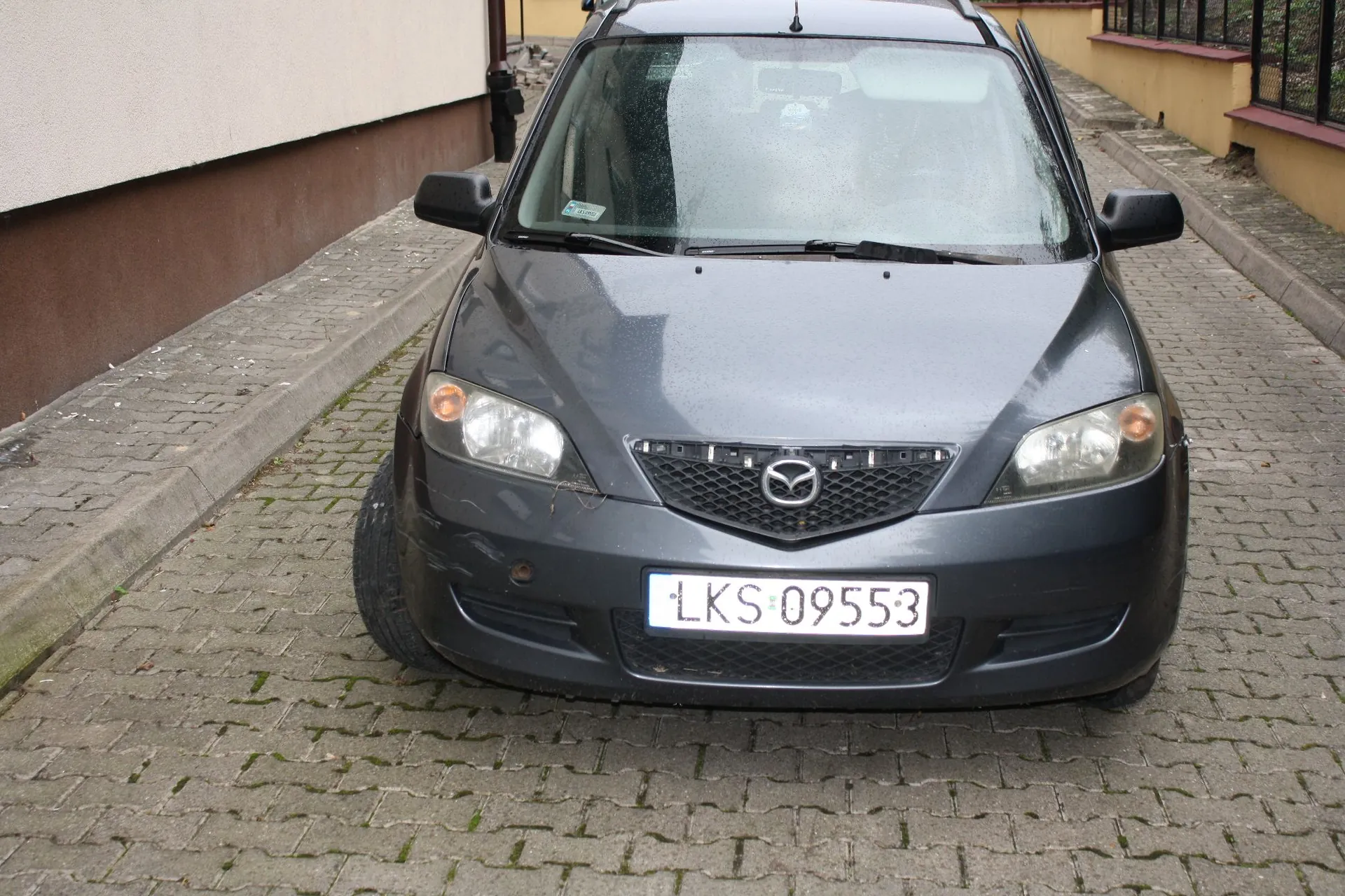 Szary samochód marki Mazda zaparkowany przodem do obiektywu na kostce brukowej. Na przodzie auta widoczna jest tablica rejestracyjna o numerze LKS 09553. Samochód znajduje się obok jasnej ściany budynku.