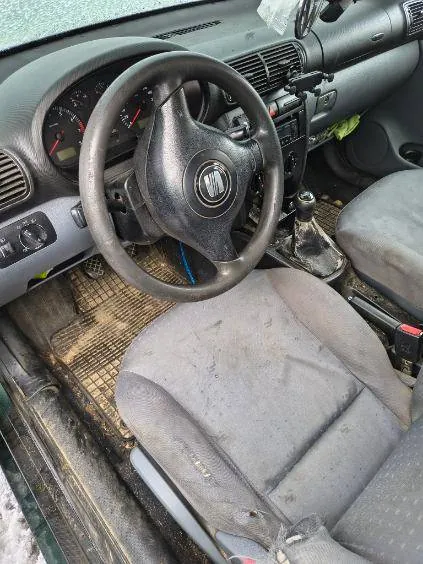 Wnętrze samochodu z czarną kierownicą z logo w środku, zegarami na desce rozdzielczej, szarymi, zabrudzonymi siedzeniami i brudnymi dywanikami podłogowymi. Widoczna dźwignia zmiany biegów oraz zielony przedmiot na desce rozdzielczej. Wnętrze wydaje się zużyte i nieczyste.