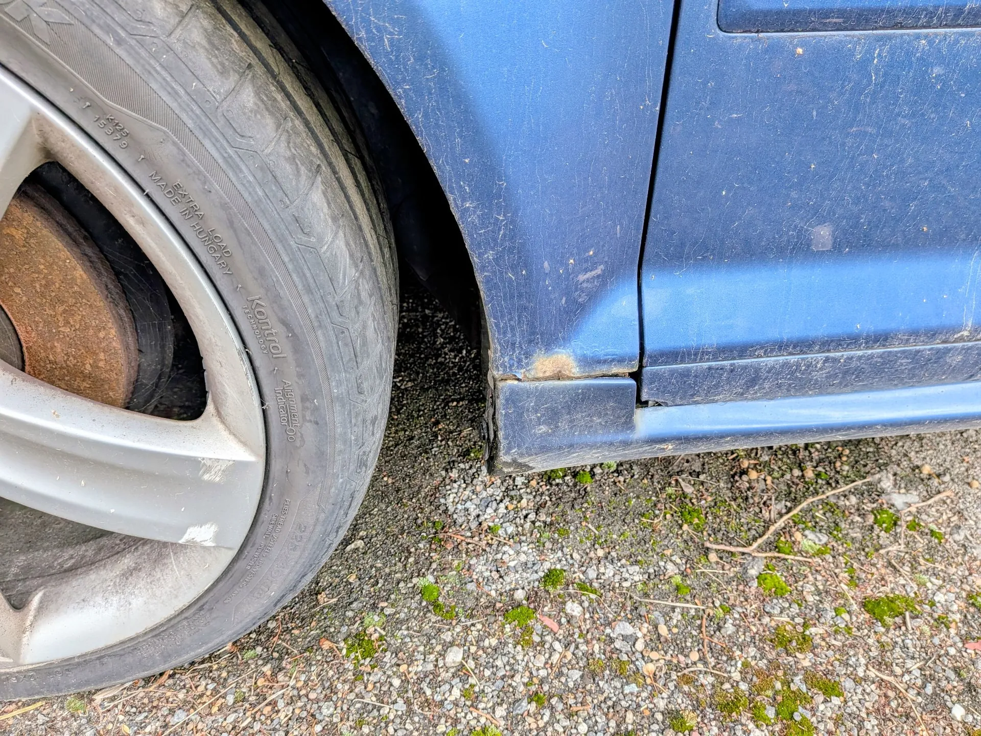 Zbliżenie na dolną część niebieskiego samochodu, pokazujące koło z felgą aluminiową oraz uszkodzony próg auta. Na krawędzi progu widoczne jest wgniecenie, odpryski lakieru i rdzawy nalot. Samochód stoi na żwirowym podłożu z kępkami mchu.