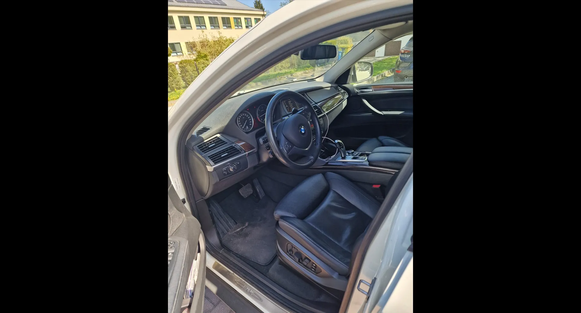 Wnętrze samochodu marki BMW widziane z otwartych drzwi kierowcy. Widoczna jest czarna skórzana tapicerka foteli, czarna kierownica z logo BMW w centrum, deska rozdzielcza z zegarami oraz konsola środkowa. Przez szybę w tle widać fragment jasnego budynku i zieleń.