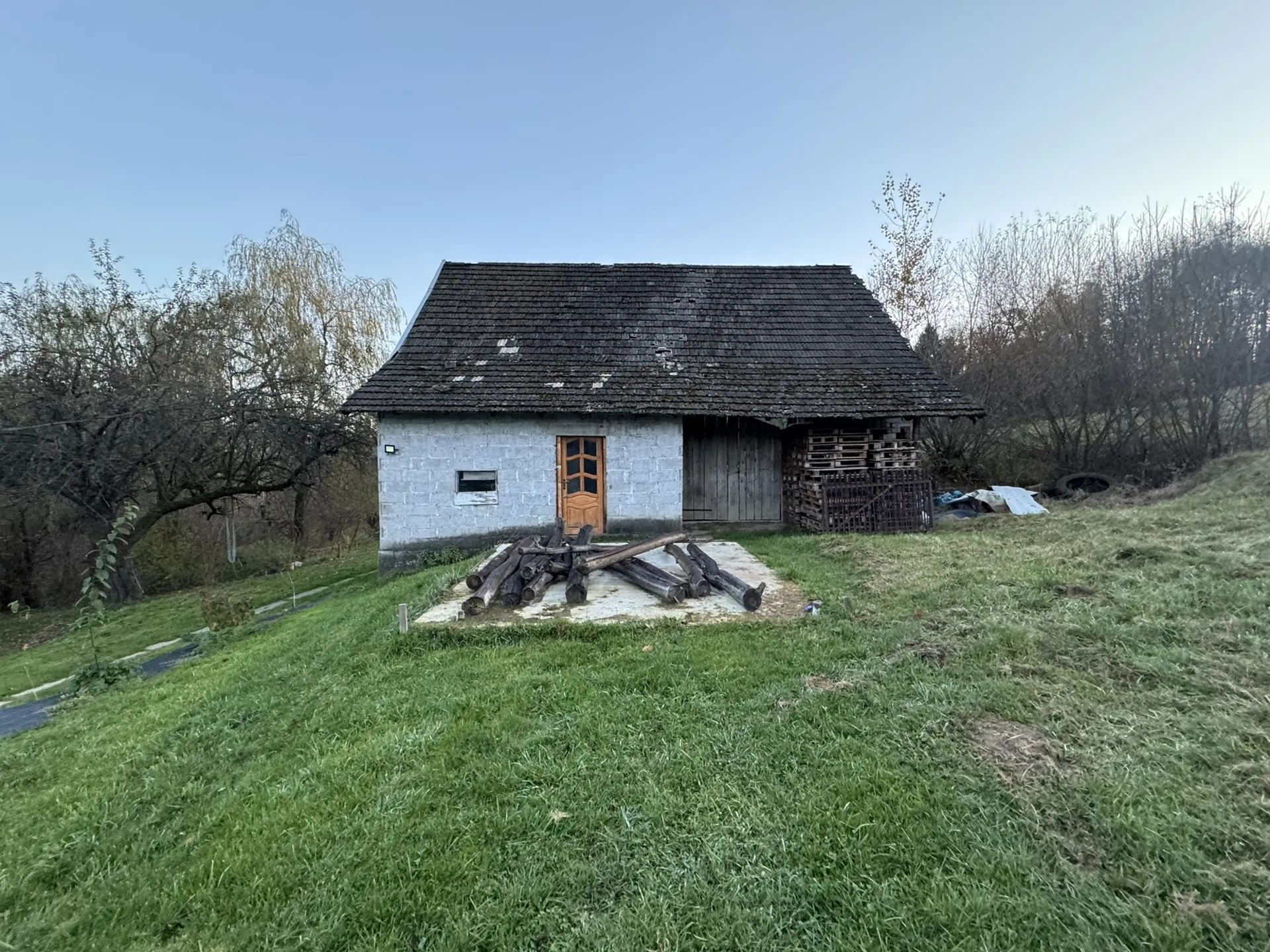 Mały dom z ciemnym dachem krytym gontem, białymi betonowymi ścianami i brązowymi drzwiami z szybkami. Przed domem na betonowej płycie leżą stosy drewna. Po prawej stronie domu znajduje się sterta palet i opon, a w tle rosną drzewa i krzewy. Całość otoczona jest zieloną trawą, a na niebie nie ma chmur.