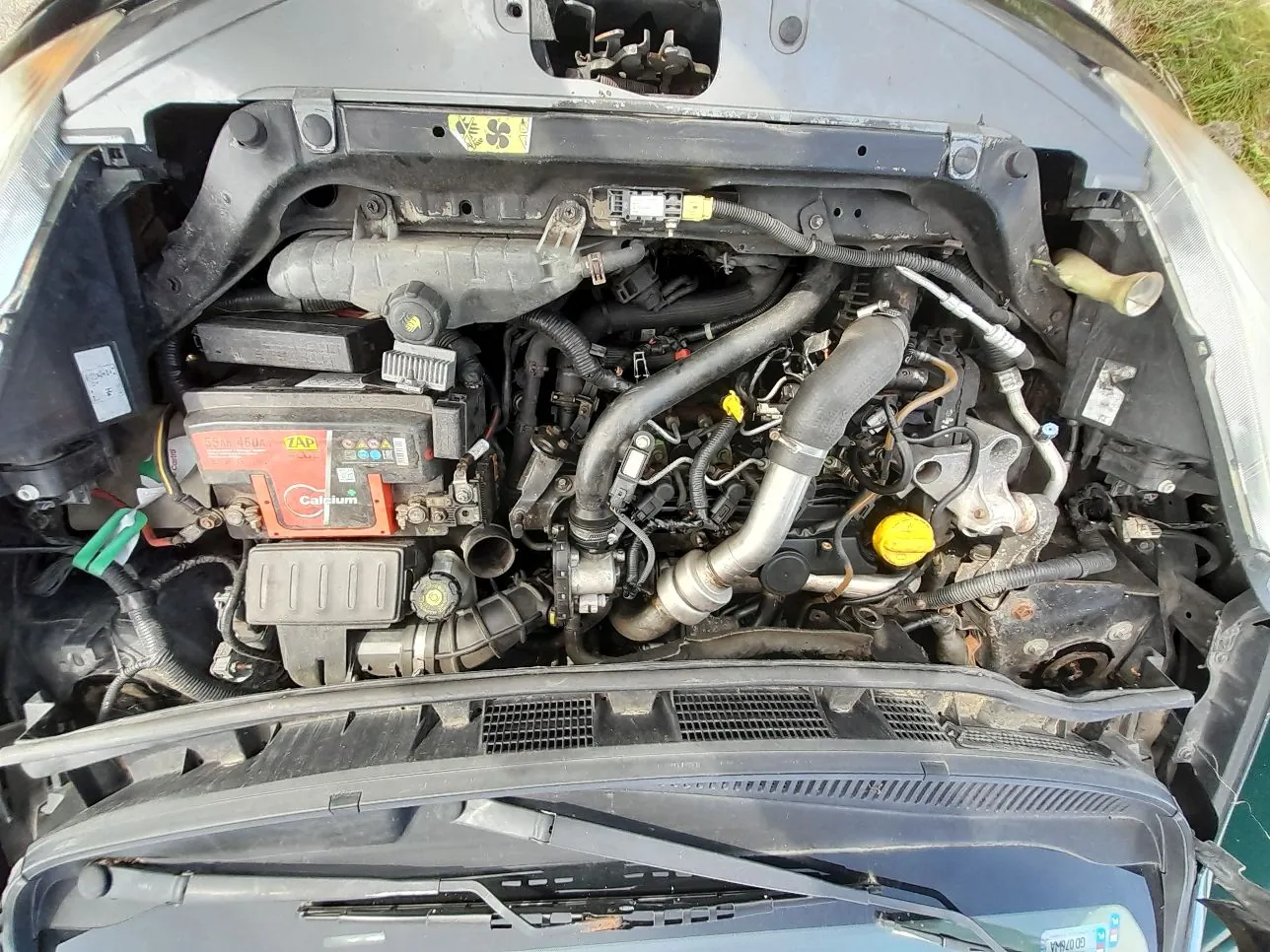 Widok z góry na komorę silnika samochodu. Widoczny jest silnik z licznymi przewodami, rurami i elementami mechanicznymi. Po lewej stronie znajduje się akumulator marki Calcium z widocznymi napisami „55Ah 450A” oraz „ZAP”. W dolnej części zdjęcia widoczny jest fragment maski i chłodnicy.
