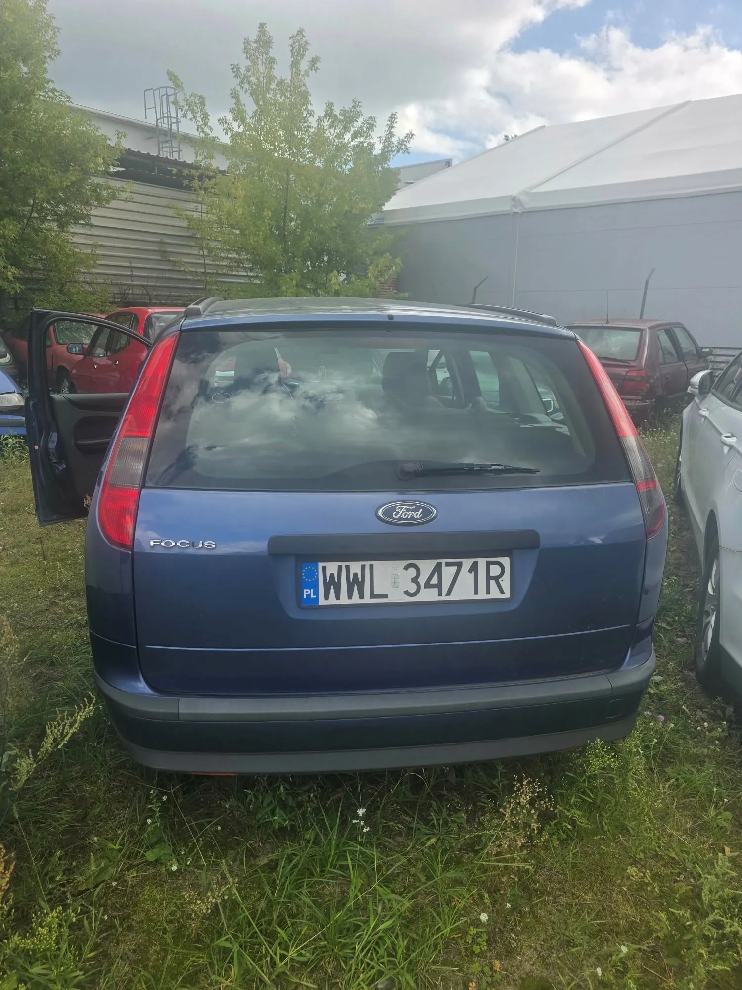 Tylny widok niebieskiego samochodu marki Ford Focus JS z polską tablicą rejestracyjną WWL 3471R. Samochód zaparkowany jest na trawie obok innych pojazdów, w tym czerwonego samochodu po lewej stronie. W tle widoczne są zielone drzewa oraz biały namiot lub hala magazynowa pod zachmurzonym niebem.
