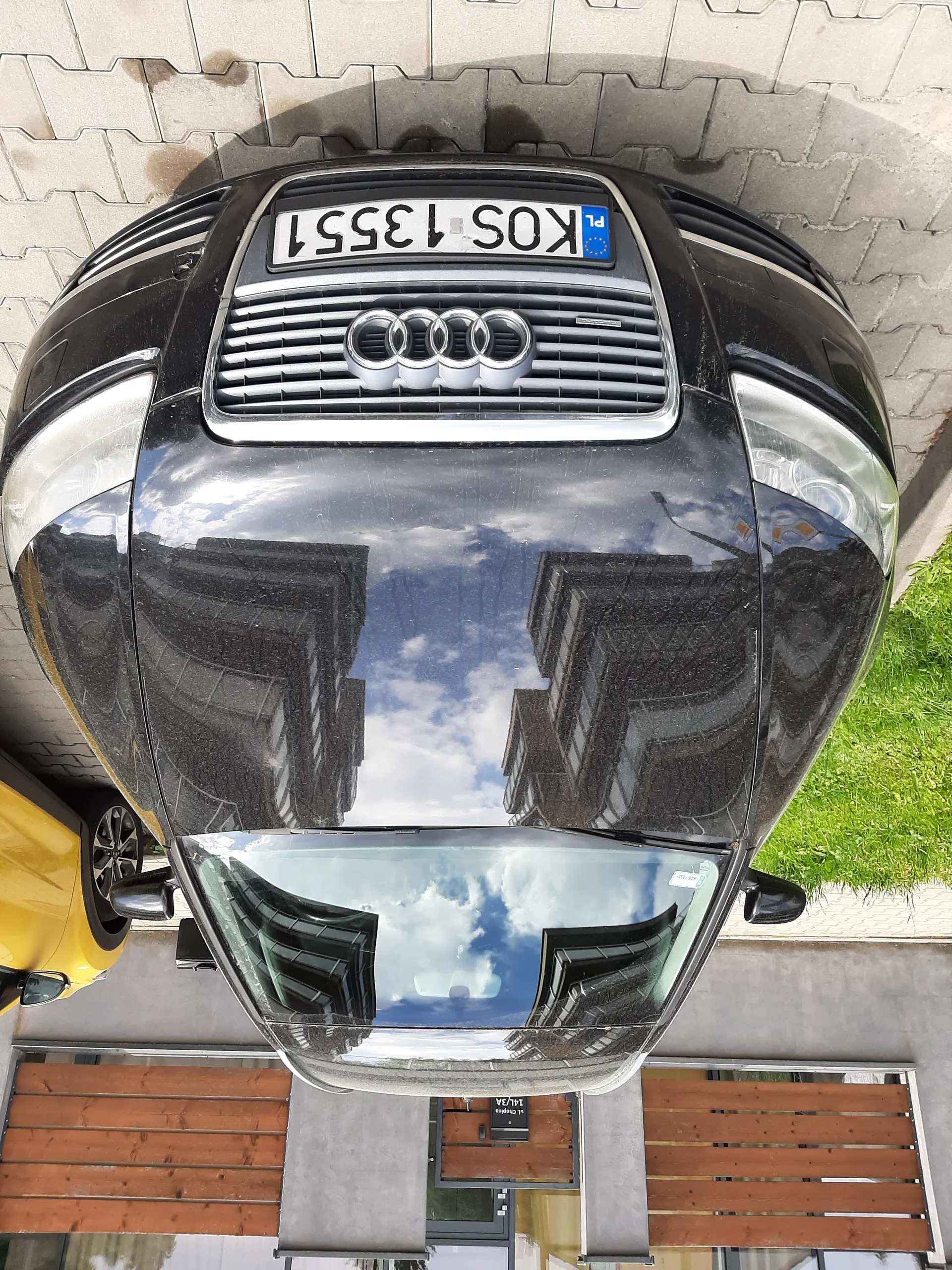 Czarny samochód marki Audi z tablicą rejestracyjną KOS 13551 zaparkowany na brukowanej powierzchni. W tle żółty samochód, budynek z drewnianymi panelami i drzwiami oraz zielona trawa. Na tablicy rejestracyjnej widoczny znak PL.