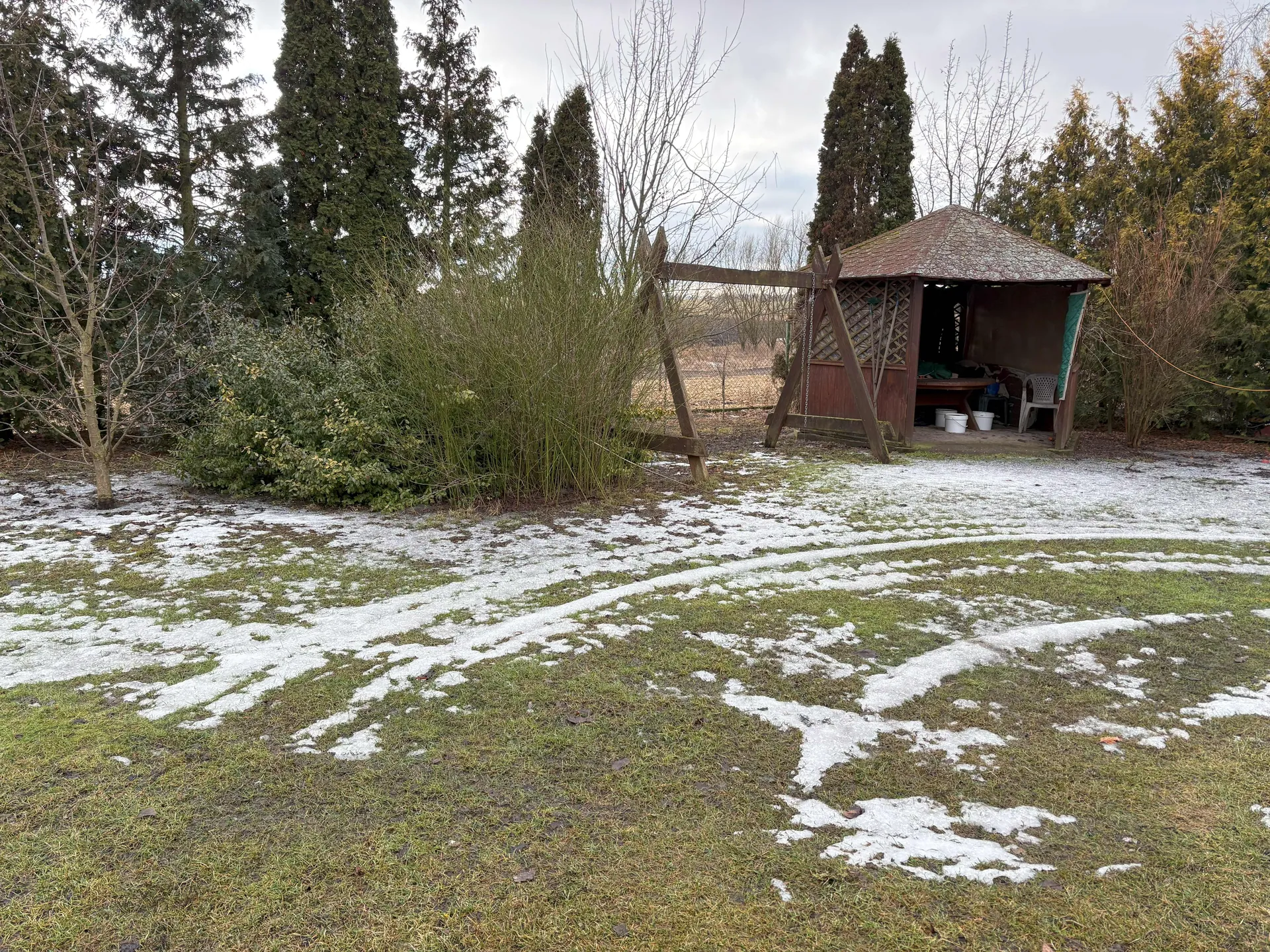 Ogrodowa scena z resztkami śniegu na trawie. Po prawej stronie drewniany pawilon z dachem krytym dachówką, pod którym znajdują się białe wiadra i plastikowe krzesło. Obok pawilonu konstrukcja z drewnianych belek przypominająca stojak na huśtawkę. W tle wysokie iglaste drzewa oraz bezlistne krzewy. Niebo jest zachmurzone.
