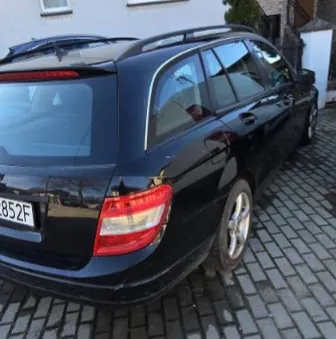 Czarny samochód typu kombi zaparkowany na brukowanej powierzchni. Widoczny tył pojazdu z tablicą rejestracyjną oznaczoną '852F', czerwonymi światłami tylnymi oraz dachowym bagażnikiem. W tle biały budynek z oknami i fragment zieleni. Samochód stoi na płaskim terenie, a horyzont jest poziomy.