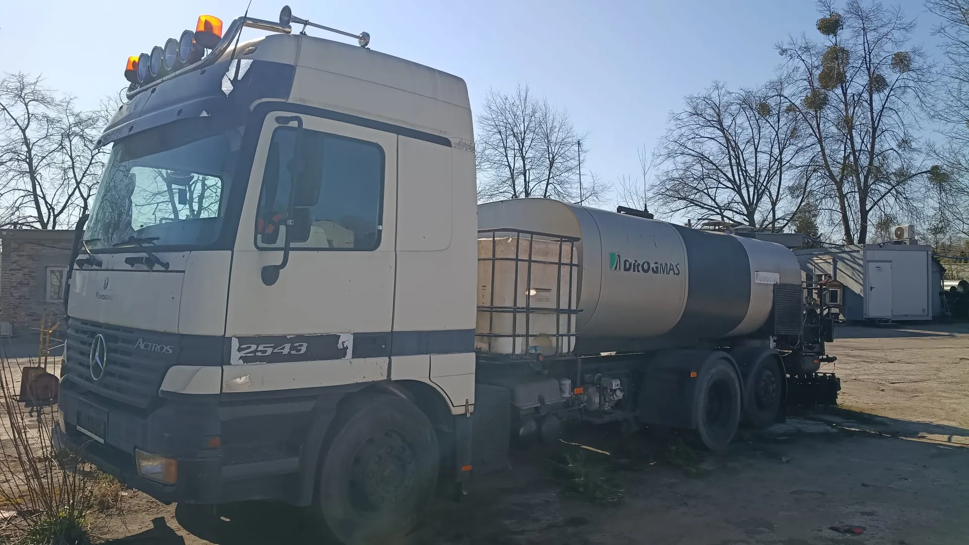 Biały ciągnik siodłowy marki Mercedes-Benz Actros z zamontowaną cysterną. Na kabinie znajdują się dodatkowe reflektory i pomarańczowe światła ostrzegawcze. Na boku kabiny widnieje napis 'ACTROS' oraz numer '2543'. Na srebrnej cysternie znajduje się zielono-niebieskie logo z napisem 'DROGMAS'. Pojazd stoi na nieutwardzonym placu na tle bezlistnych drzew i jasnego nieba.