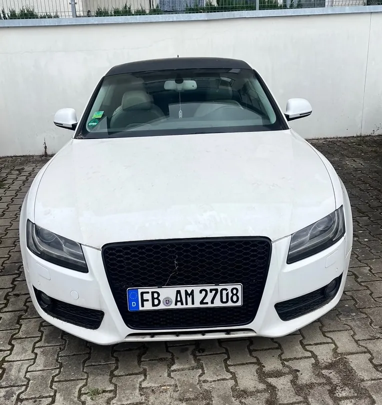 Biały samochód osobowy typu kabriolet zaparkowany przodem do widza na kostce brukowej. Pojazd ma czarny grill i niemiecką tablicę rejestracyjną o treści FB AM 2708. W tle widoczna jest biała ściana i fragment ogrodzenia.