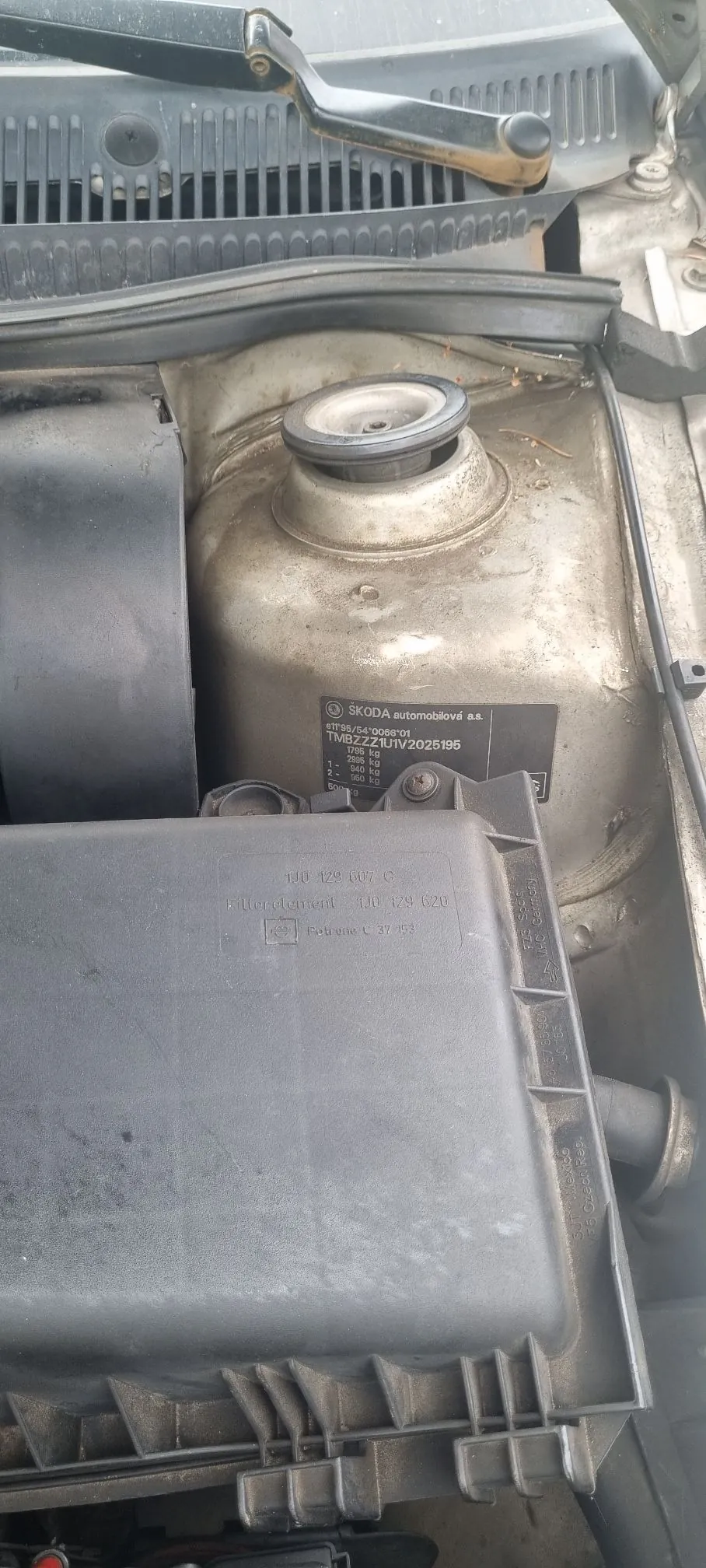 Zbliżenie na komorę silnika samochodu. Widoczny jest metalowy element nadwozia z okrągłym mocowaniem amortyzatora oraz plastikowy zbiornik wyrównawczy z tekstem: 'SKODA automobilové a.s.', 'TMBZ-ZAUTIV2025195', '1795 kg', '1. 2000 kg', '2. 940 kg', '3. 500 kg'. Poniżej znajduje się szary, plastikowy pojemnik z oznaczeniami '1K0 129 007 G' oraz '1K0 129 020'.
