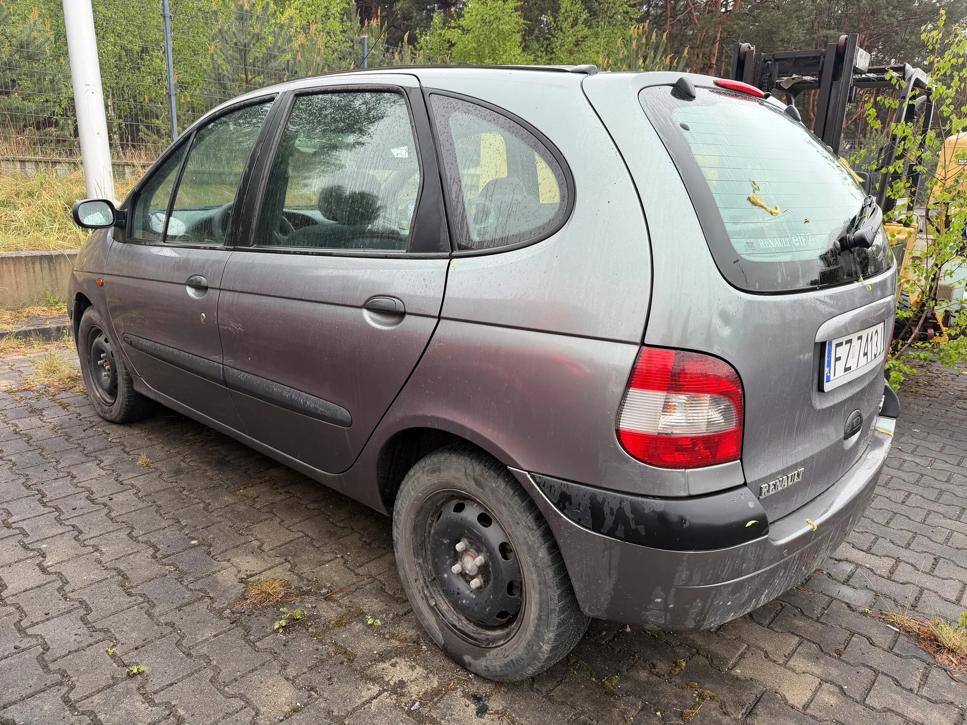 Szary samochód marki Renault, widok z tyłu i boku. Pojazd stoi na brukowanej powierzchni, pokrytej plamami mchu i suchą trawą. Na tylnym szybie widoczny napis 'RENAULT' oraz tablica rejestracyjna 'FZ 7413'. Samochód ma brudną karoserię z kroplami wody, czarne felgi i czerwone światła tylne. W tle: metalowe ogrodzenie, zielone drzewa, żółty pojazd budowlany oraz słup. Na szybie tylnej widoczne są żółte plamy.