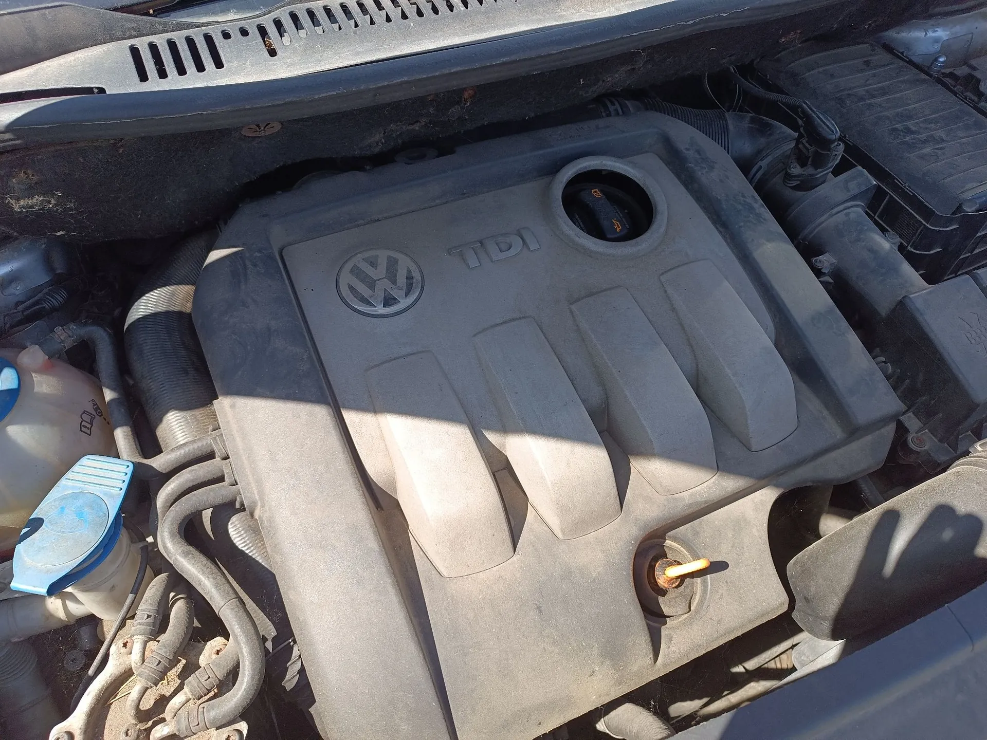 Zbliżenie na silnik samochodu z szarą plastikową osłoną. Na osłonie widoczne jest logo marki Volkswagen oraz napis 'TDI'. W górnej części znajduje się wlew oleju, a w dolnej części bagnet do pomiaru poziomu oleju. Po lewej stronie widoczny jest zbiornik z niebieską zakrętką oraz przewody.
