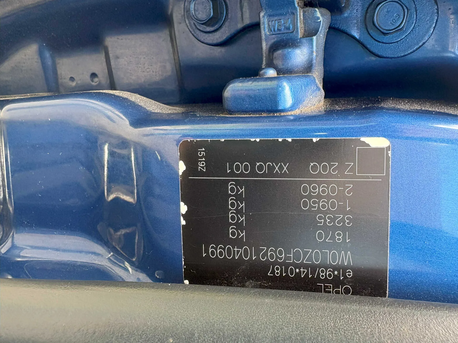 Czarna tabliczka z białym tekstem zamontowana na niebieskim metalowym elemencie pojazdu. Widoczne napisy: OPEL, e1*98/14*0187, W0LOZCF6921040991, 1870 kg, 3235 kg, 1-0950 kg, 2-0960 kg, Z 20Q, XXJQ 001, 1519Z. Po lewej stronie tabliczki metalowe elementy z gwintowanymi śrubami i zawiasem.