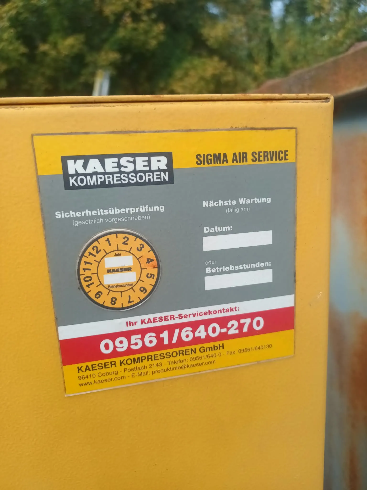 Żółta obudowa urządzenia z naklejką serwisową. Na naklejce widnieje napis 'KAESER KOMPRESSOREN' oraz 'SIGMA AIR SERVICE'. Poniżej znajduje się sekcja 'Sicherheitsüberprüfung (gesetzlich vorgeschrieben)' z okrągłym schematem miesięcy roku oraz sekcja 'Nächste Wartung (fällig am)' z polami do wpisania daty lub godzin pracy (Betriebsstunden). Na dole widnieje czerwony pasek z numerem telefonu '09561/640-270' oraz dane firmy 'KAESER KOMPRESSOREN GmbH' z adresem w Coburg i adresem strony internetowej www.kaeser.com.